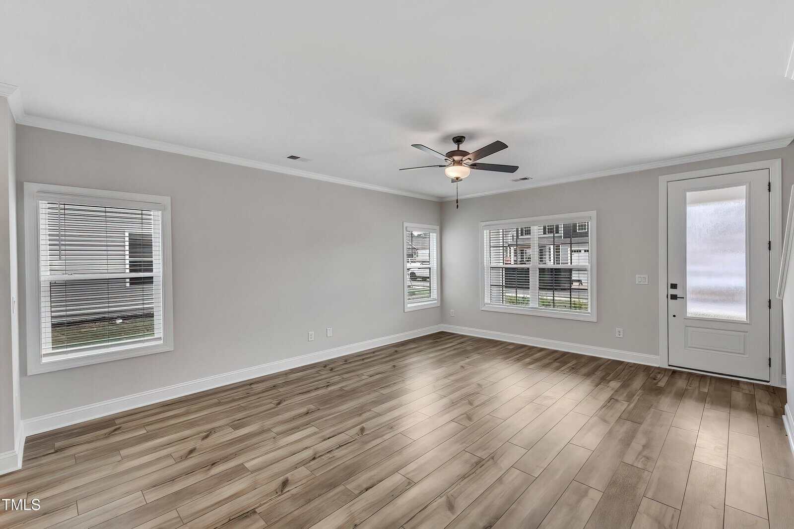 Empty living room with light gray walls, wood-look tile flooring, three large windows with blinds, a ceiling fan with a light fixture, and a white door with frosted glass panel.