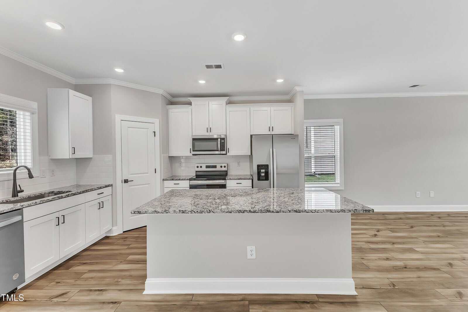Modern kitchen with white cabinets, stainless steel appliances, granite countertops, island with outlet, wood-look flooring, and recessed lighting. Door and windows provide natural light; open layout leads to living area.