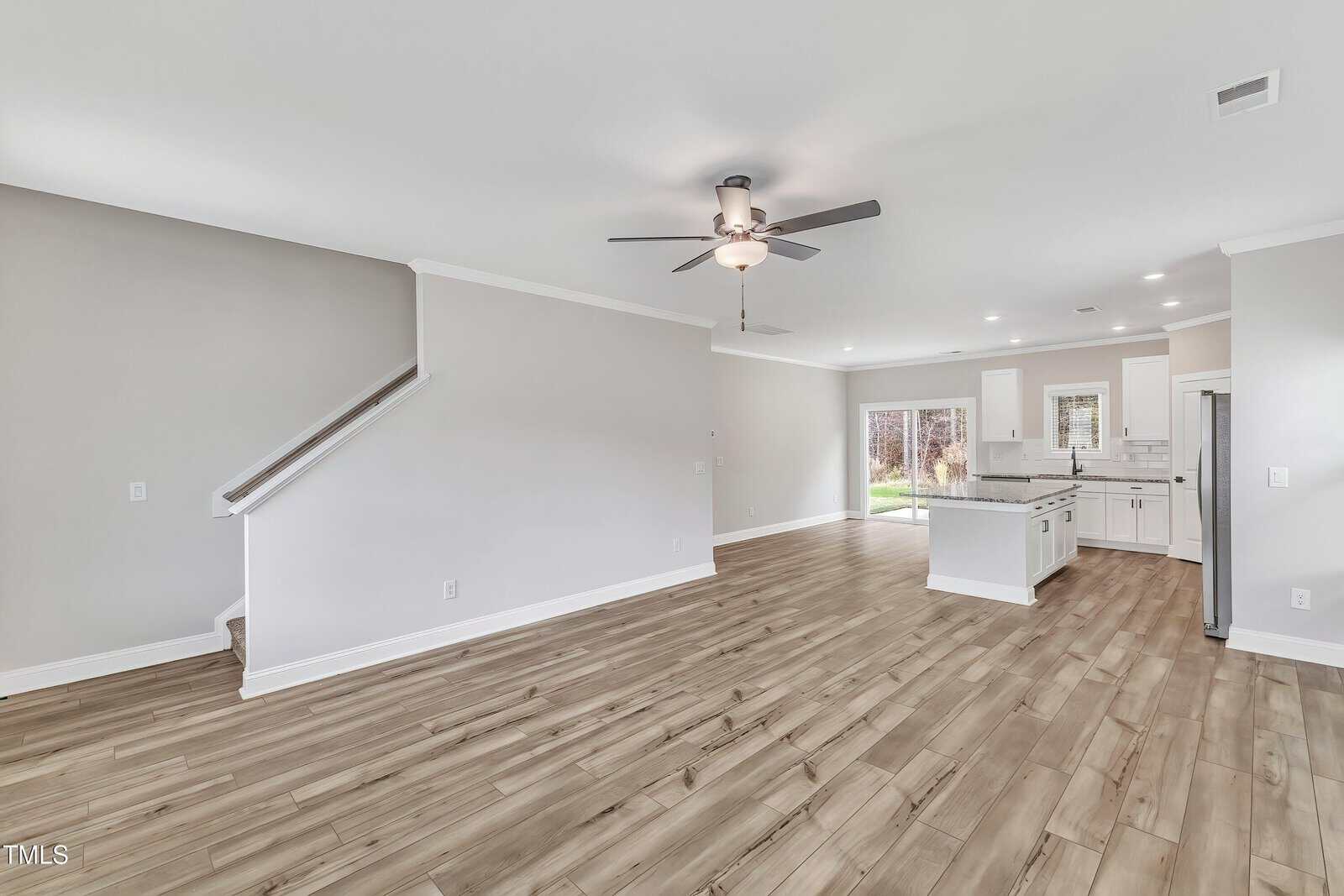 Open-concept living area with light wood flooring, neutral walls, ceiling fan, and recessed lighting; features a staircase on the left and a kitchen with white cabinets, island, and stainless steel appliances on the right.