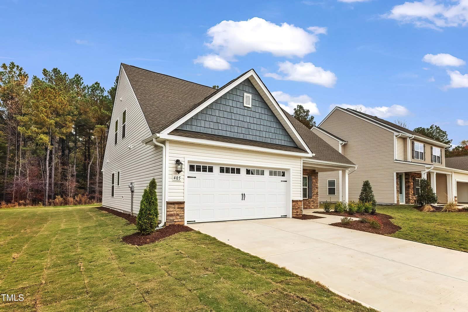 A two-story suburban house with gray siding, white trim, and a double garage door. The driveway and lawn are neatly maintained, and the house is surrounded by trees and neighboring homes. The sky is partly cloudy.