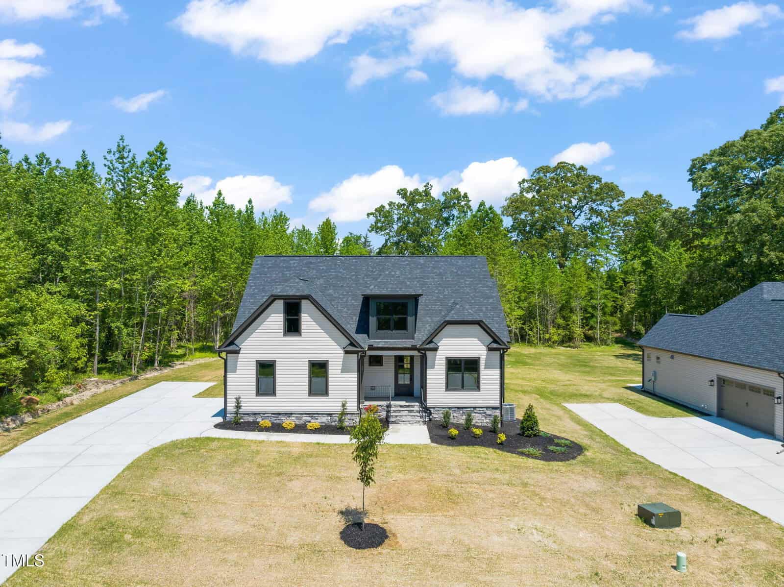 A two-story house with light-colored siding, dark roof, and black trim sits on a spacious lot with a large driveway, young landscaping, and surrounded by trees under a partly cloudy sky.