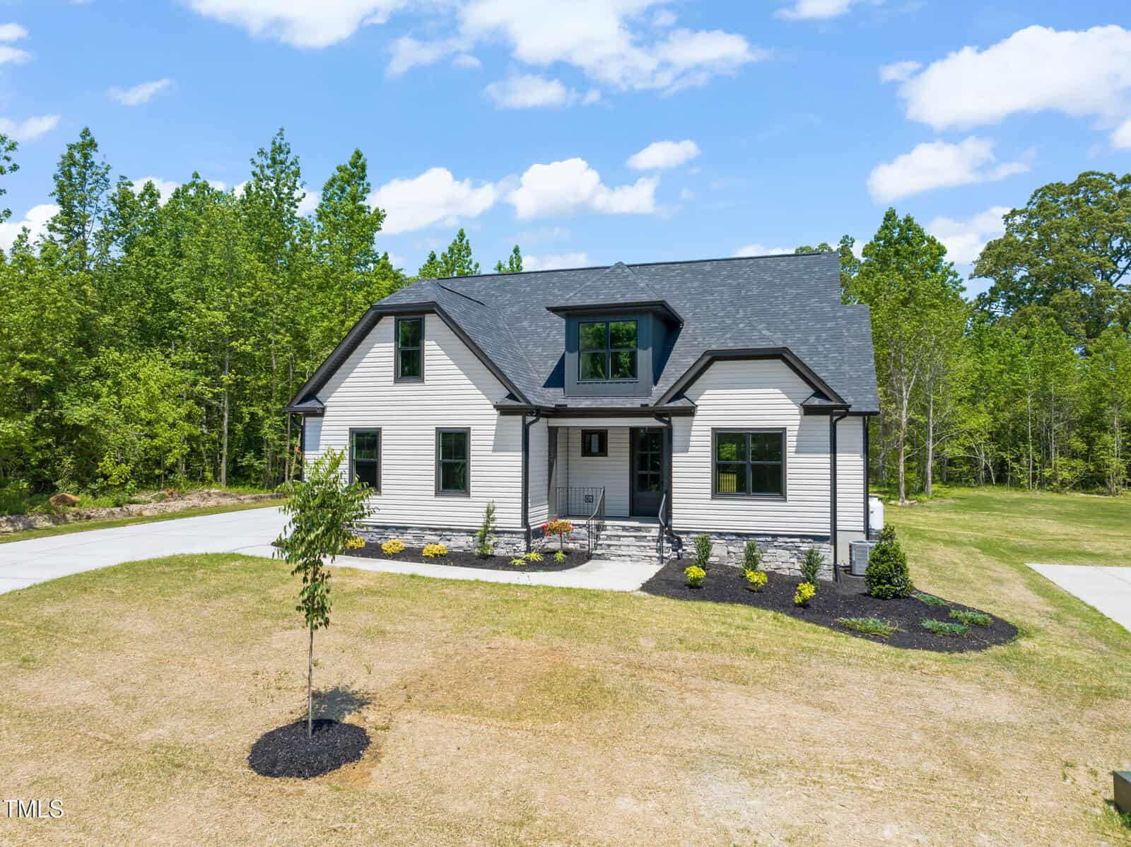 A two-story house with light-colored siding, dark trim, and a dark roof sits on a large lot with a curved driveway, small front shrubs, and surrounded by green trees under a blue sky with scattered clouds.