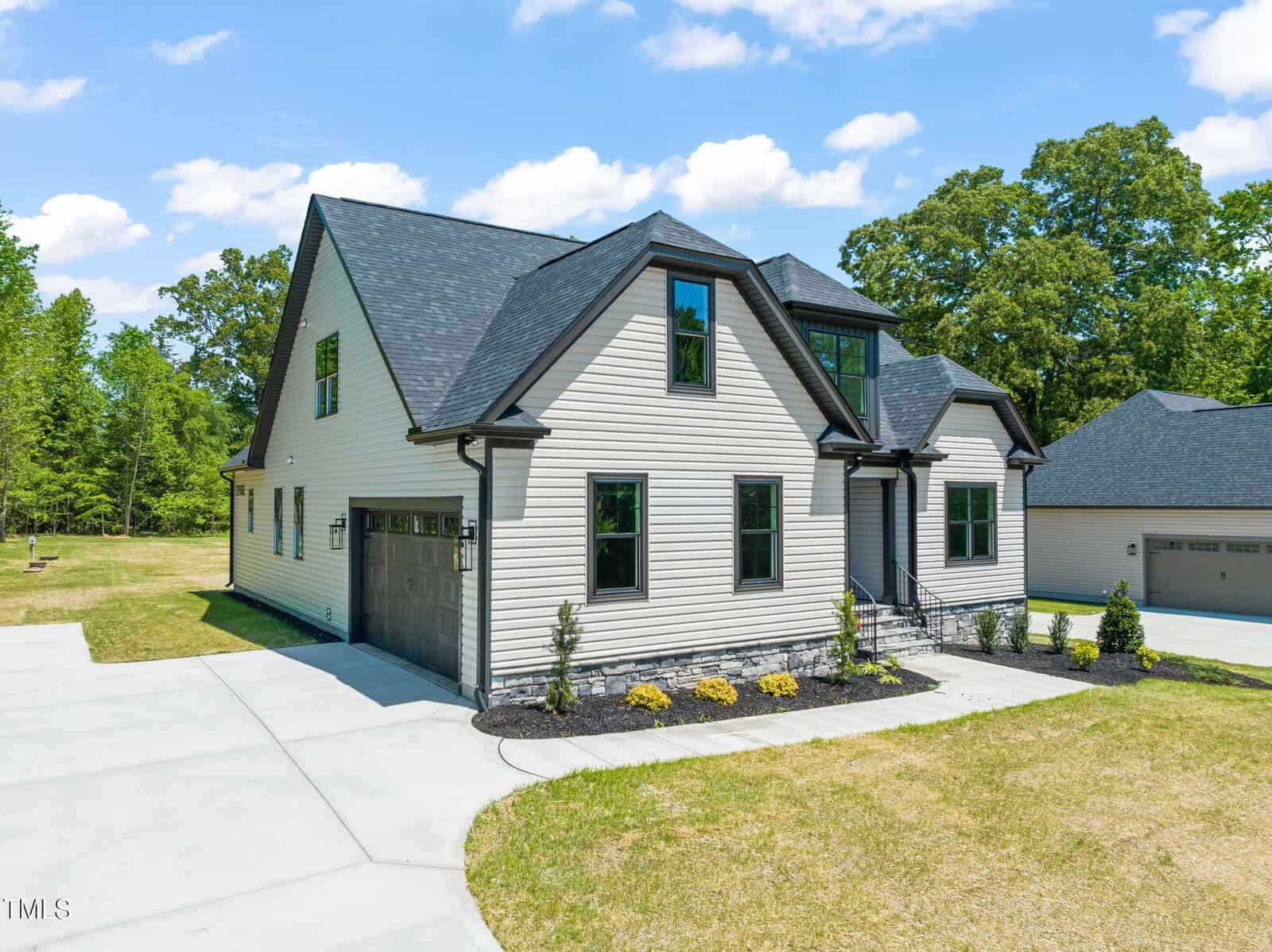 A modern two-story house with light-colored siding, dark roof shingles, a two-car garage, black-trimmed windows, and a small landscaped area in front, set on a spacious lot with a concrete driveway.