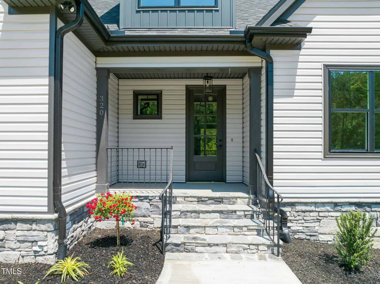 A front view of a house entrance with steps leading to a black door, black railings, stone and siding exterior, house number 320, and small landscaped plants and flowers by the walkway.