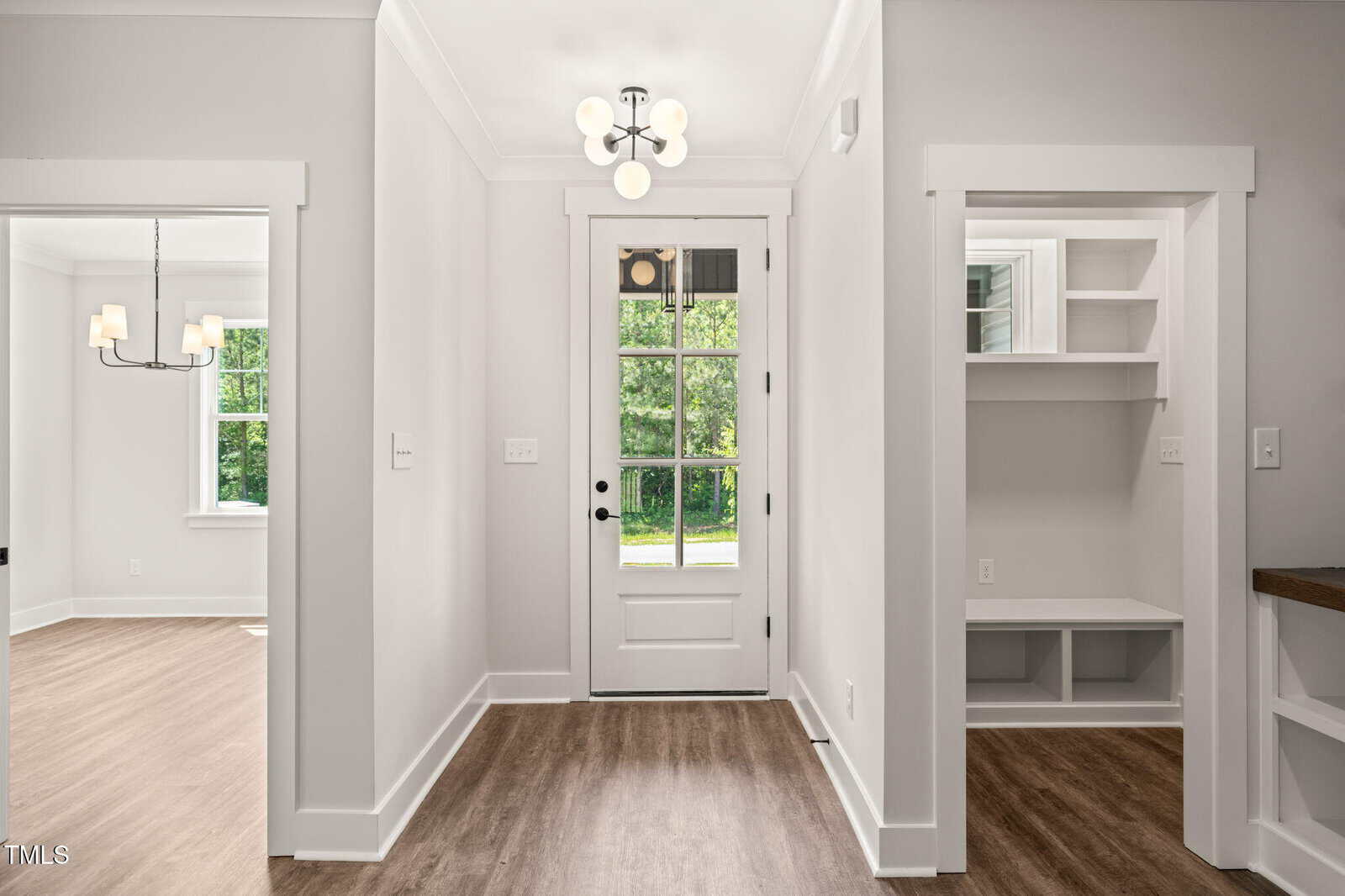 A bright entryway with a glass-paneled door, light walls, and wood flooring. To the left is a room with a window and chandelier, and to the right is a built-in storage nook with shelves and a bench.