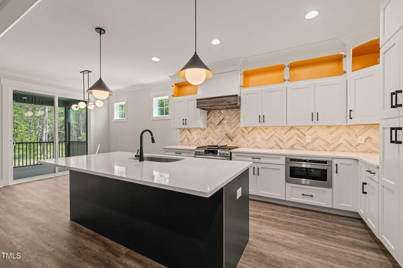 Modern kitchen with white cabinets, wood floors, black island with white countertop, three pendant lights, stainless steel appliances, herringbone backsplash, and sliding doors leading to a covered outdoor patio.
