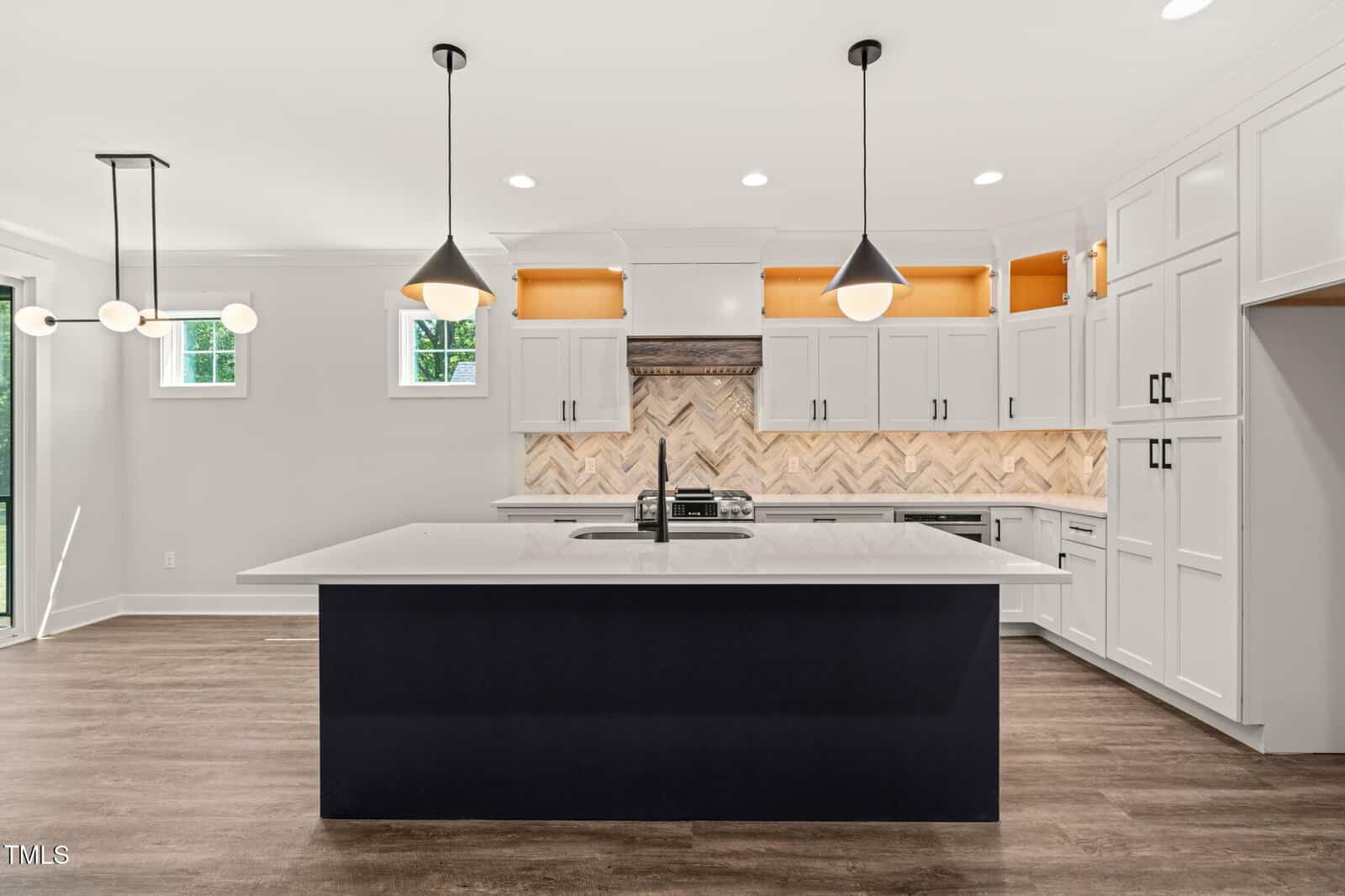 Modern kitchen with a large black island, white countertops, white cabinets, pendant lights, herringbone backsplash, wood floors, and built-in lighting in cabinet niches. Two small windows provide natural light.