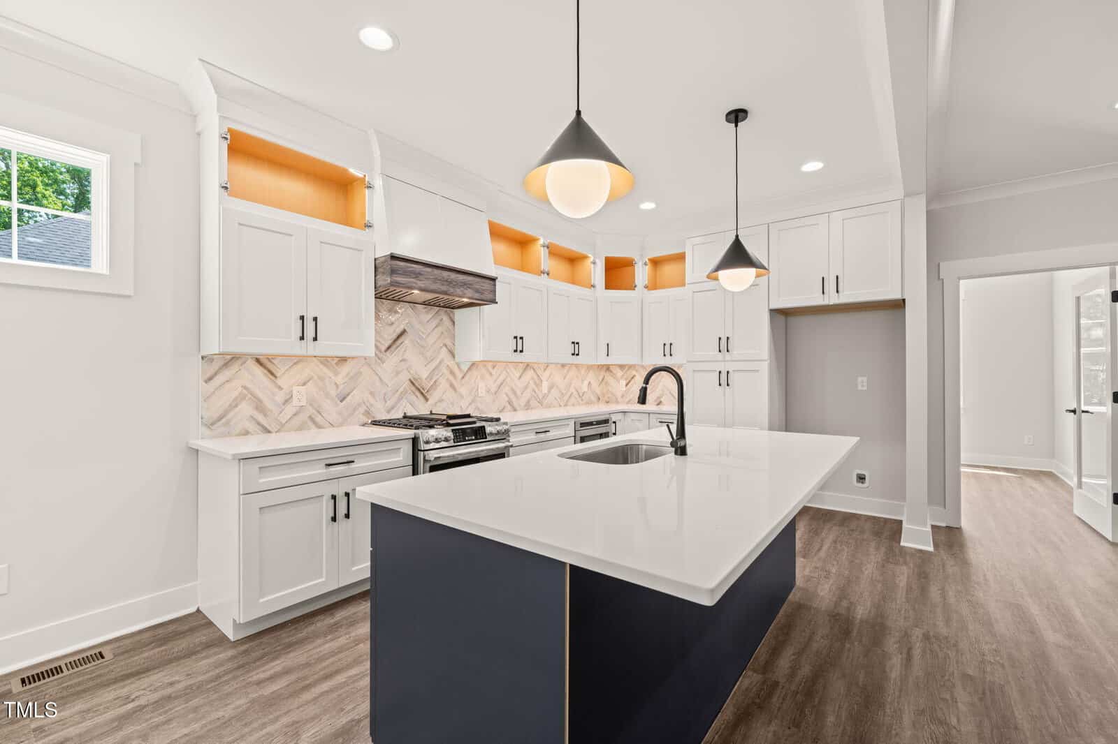 Modern kitchen with white cabinets, a large island with a black base and white countertop, stainless steel stove, herringbone backsplash, pendant lights, and wood floors. Doorways lead to adjacent rooms.