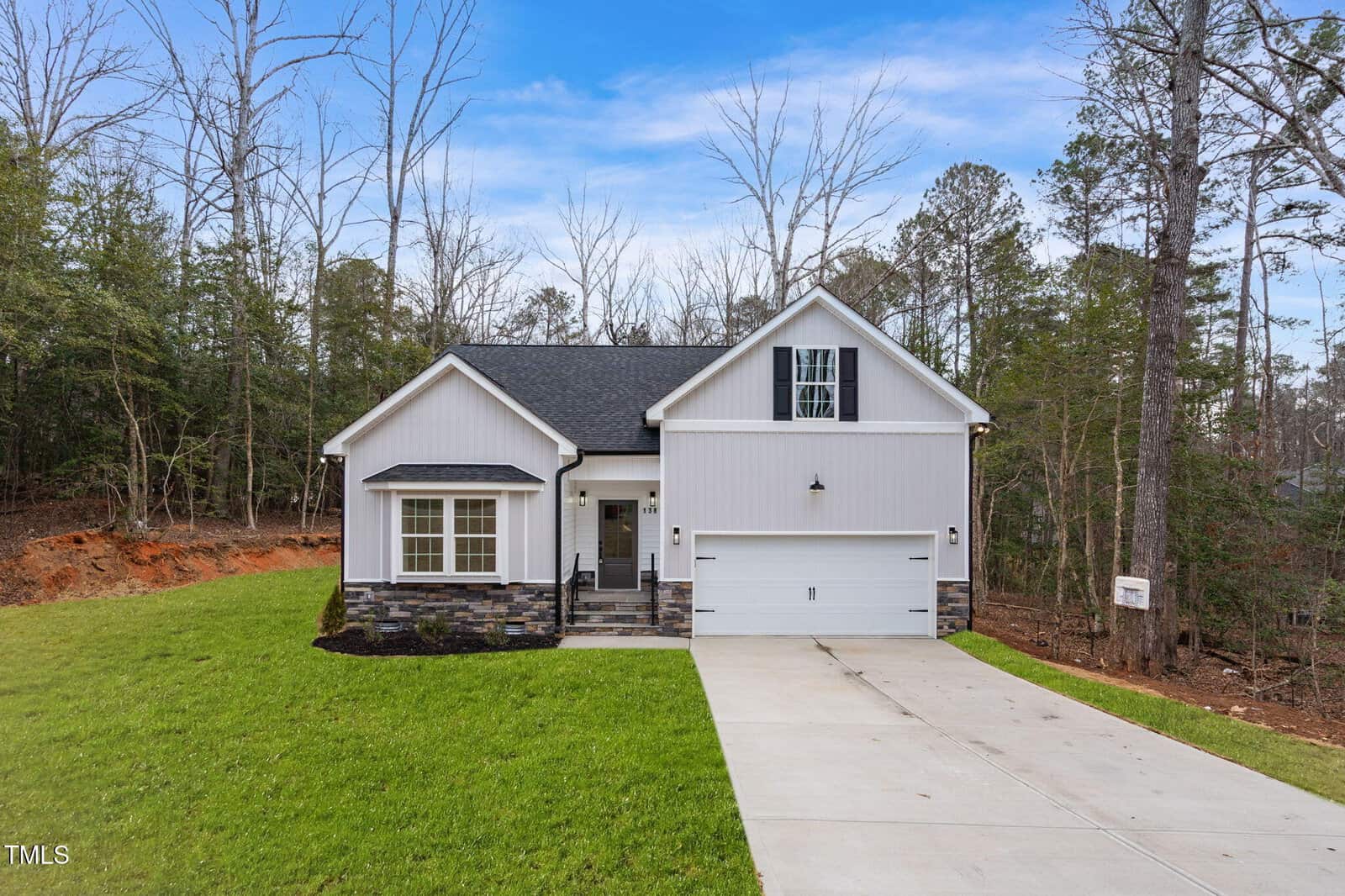 A modern, single-story house with gray siding and stone accents, attached two-car garage, bay window, concrete driveway, and green lawn, surrounded by trees under a blue sky.