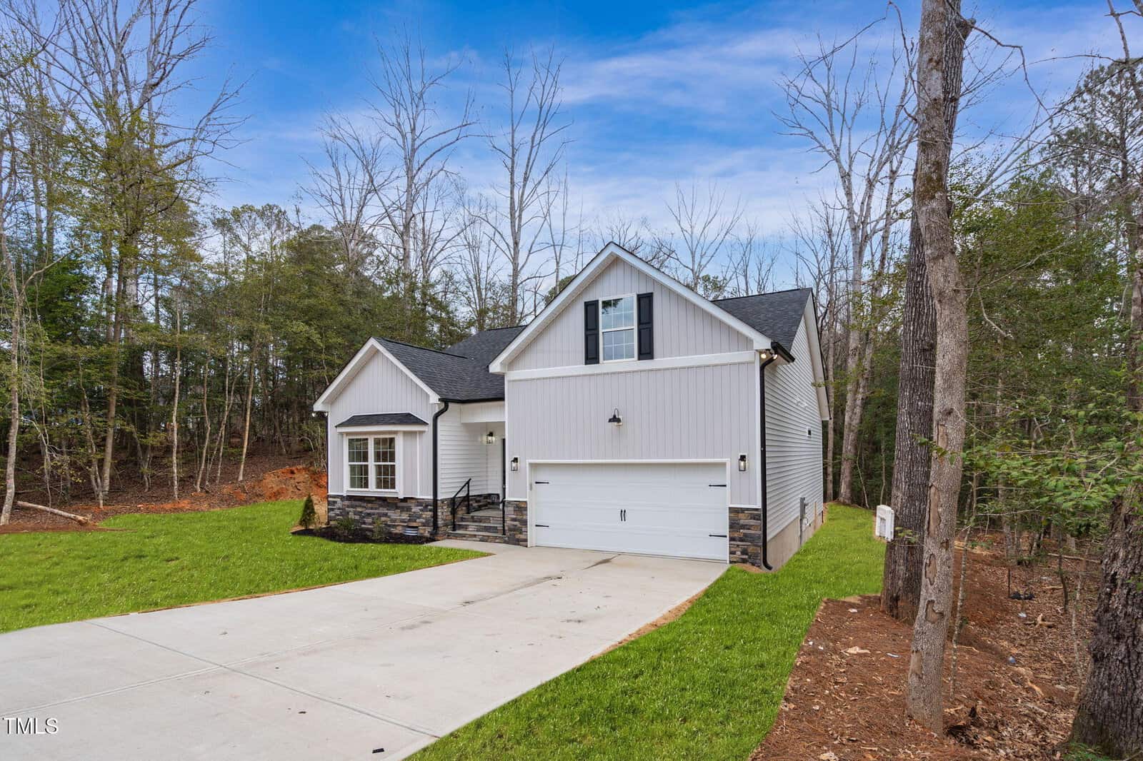 A modern single-story house with gray siding, stone accents, a double garage, and a concrete driveway. The home is surrounded by grass, trees, and a partially wooded yard under a blue sky.