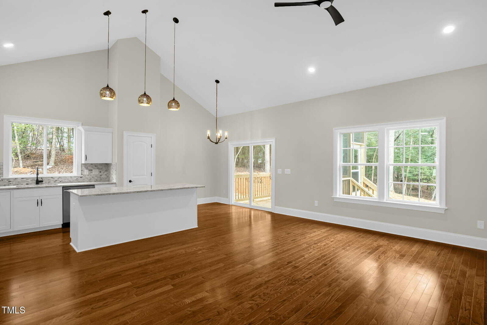 Open concept living area with hardwood floors, a white kitchen with an island and pendant lights, large windows, a chandelier over the dining space, and a ceiling fan, with views of a wooden deck and trees outside.