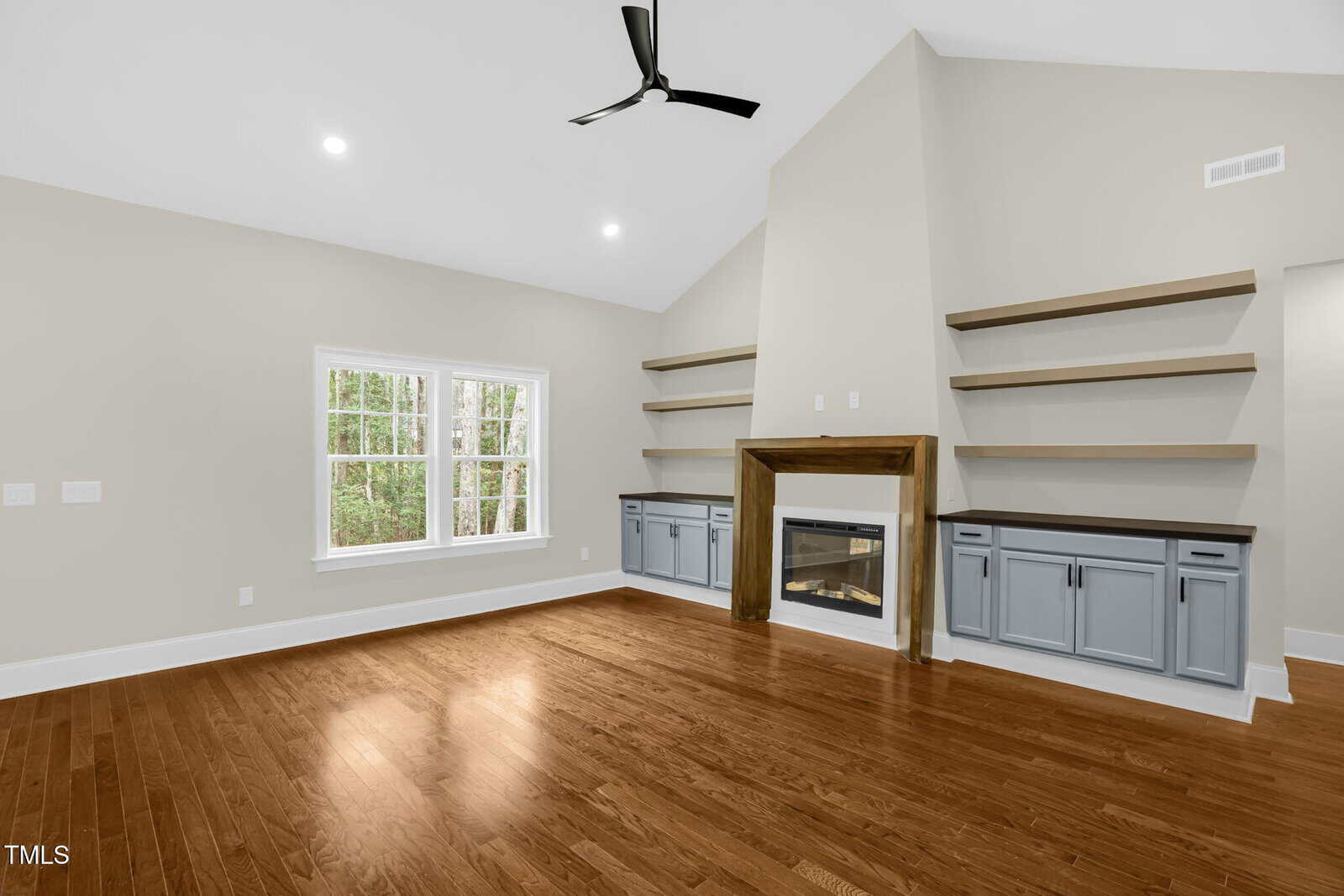 A spacious living room with hardwood floors, a ceiling fan, a fireplace with a wooden mantel, built-in shelves and cabinets, and a large window letting in natural light.