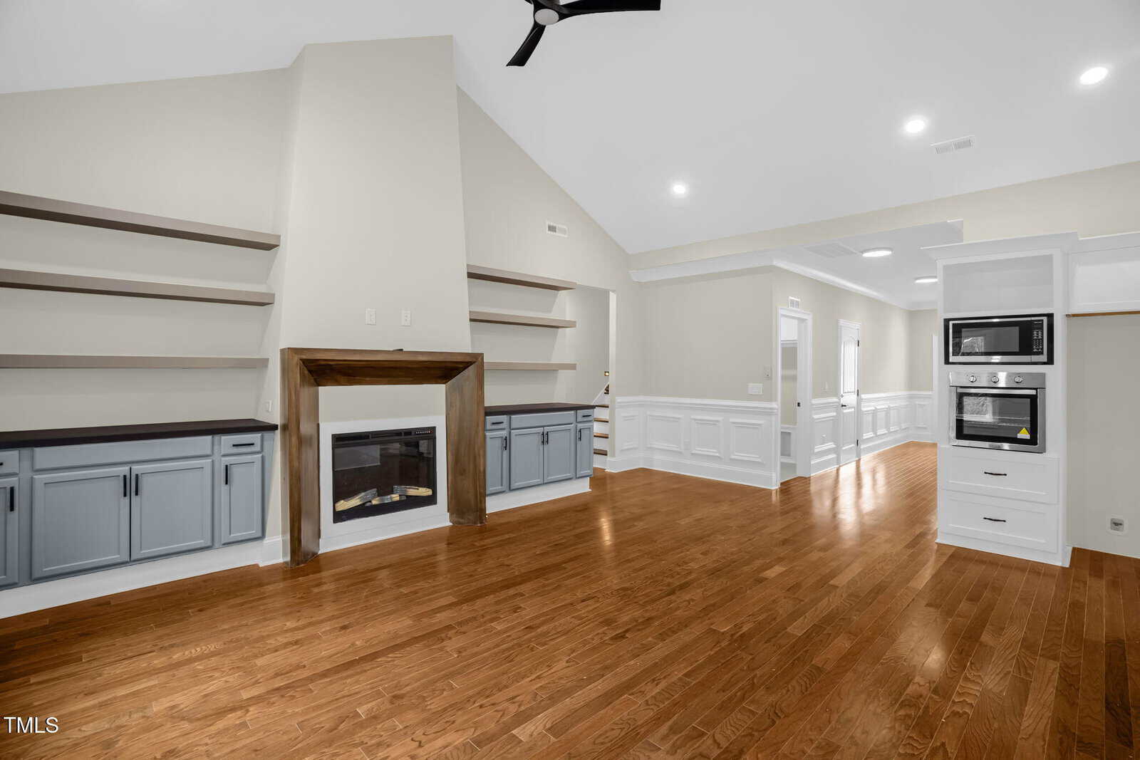 A spacious living room with hardwood floors, built-in shelves and cabinets, a fireplace with a wooden mantel, neutral walls, and a view into a white kitchen area with built-in microwave and oven.