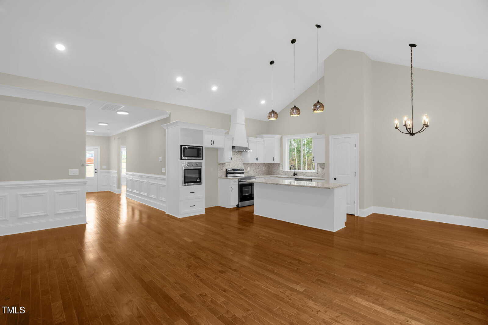Modern kitchen with white cabinets, stainless steel appliances, an island with pendant lights, and adjacent dining area. Hardwood floors and neutral walls throughout. White door and windows let in natural light.