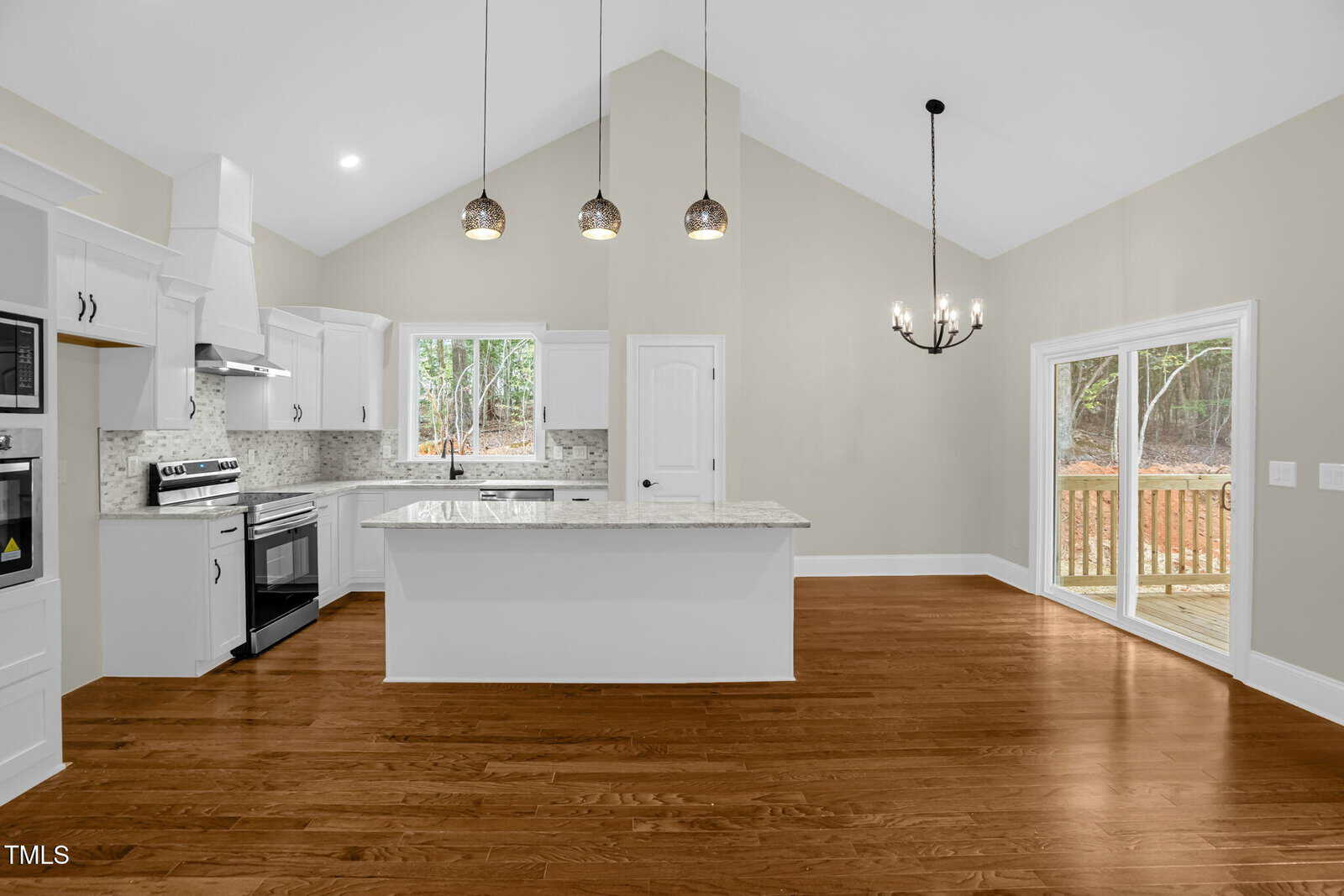 Modern kitchen with white cabinets, stainless steel appliances, a marble island, pendant lights, hardwood floors, and an adjacent dining area with a black chandelier and sliding glass doors leading to a wooden deck.