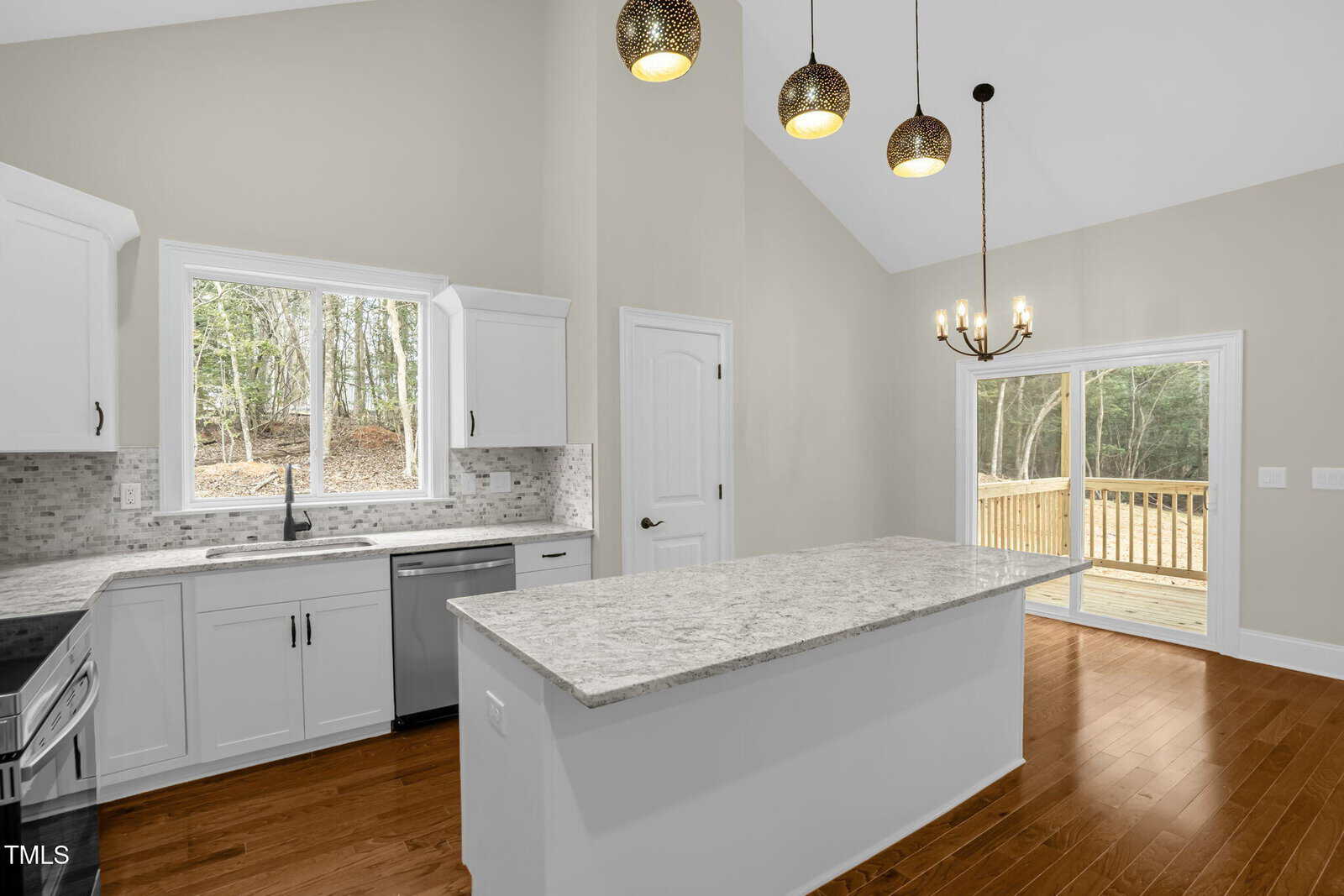 Modern kitchen with white cabinets, light granite countertops, stainless steel appliances, an island, pendant lights, wood flooring, a window over the sink, and sliding glass doors leading to a deck with a wooded view.