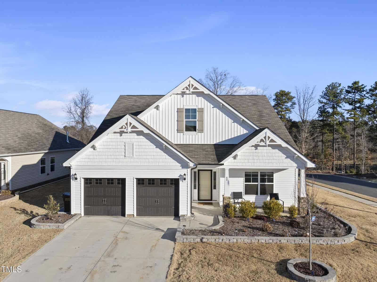 A modern single-story house with white siding, dark brown double garage doors, a gabled roof, and a front porch. The yard has minimal landscaping with bushes and mulch beds, and the driveway is concrete.
