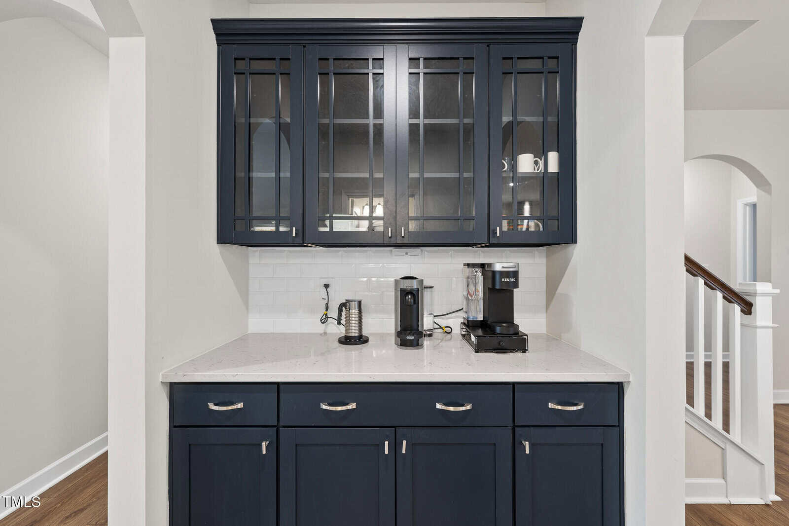 A coffee bar area with navy cabinets, a white countertop, a coffee maker, a kettle, a frother, and glass-front upper cabinets against a white subway tile backsplash. Stairs are visible to the right.
