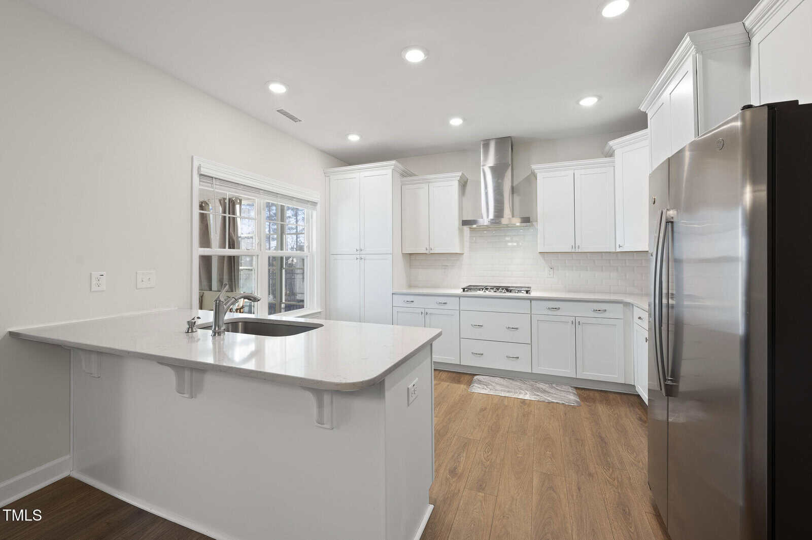 Modern kitchen with white cabinets, stainless steel appliances, a range hood, tile backsplash, and a large island with a sink. Wood flooring and recessed lighting create a clean, bright atmosphere.