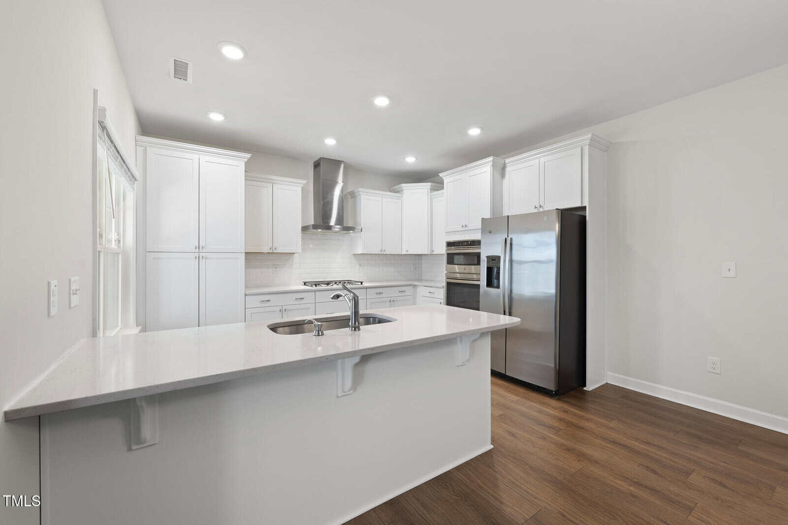 Modern kitchen with white cabinets, stainless steel appliances, a center island with sink, and wood flooring. Recessed ceiling lights provide bright illumination. The walls are light-colored, giving a clean appearance.