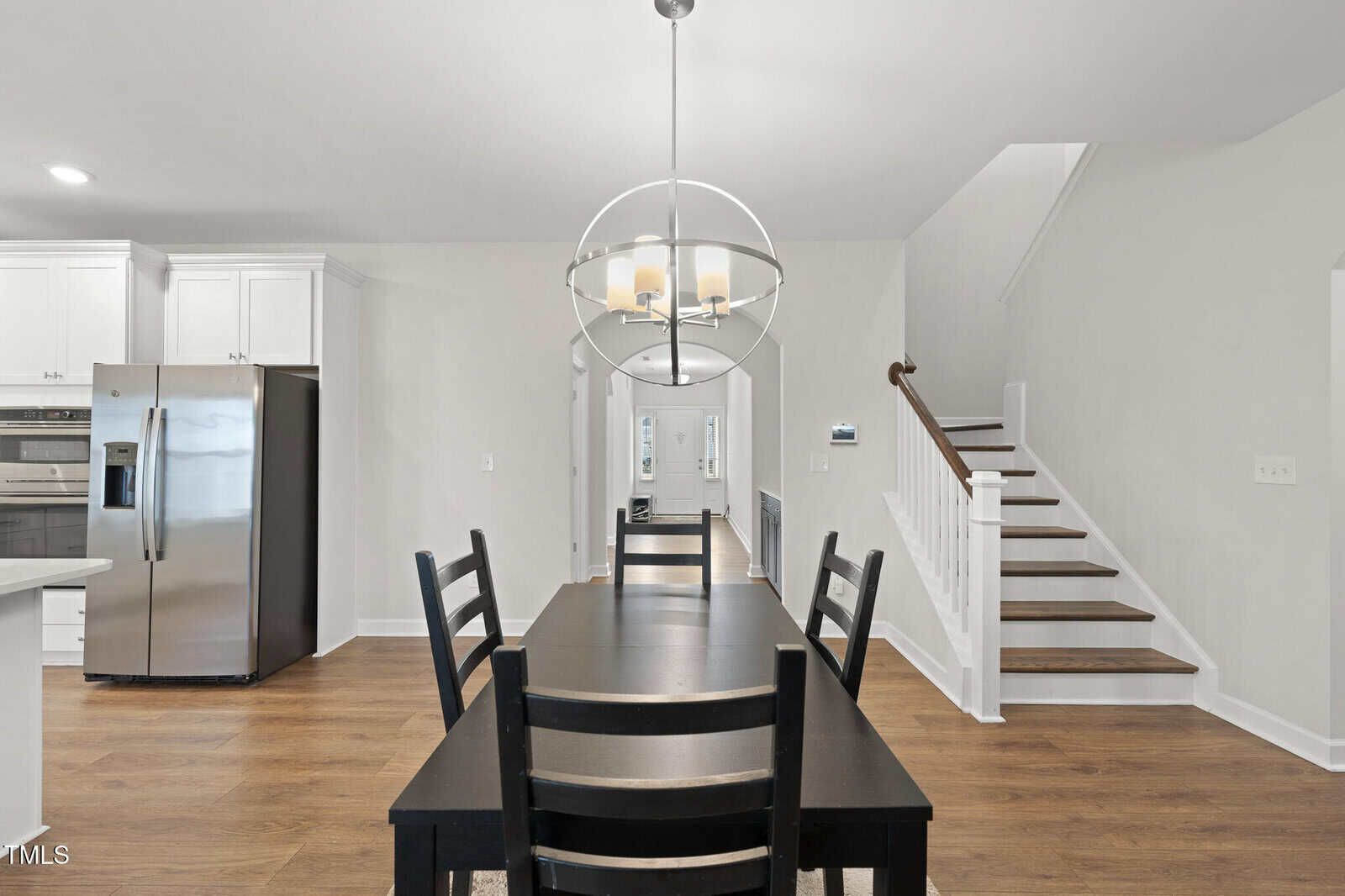 A modern kitchen and dining area with a black dining table and chairs, stainless steel refrigerator, white cabinets, wooden floor, and a staircase leading upstairs. A spherical chandelier hangs above the table.