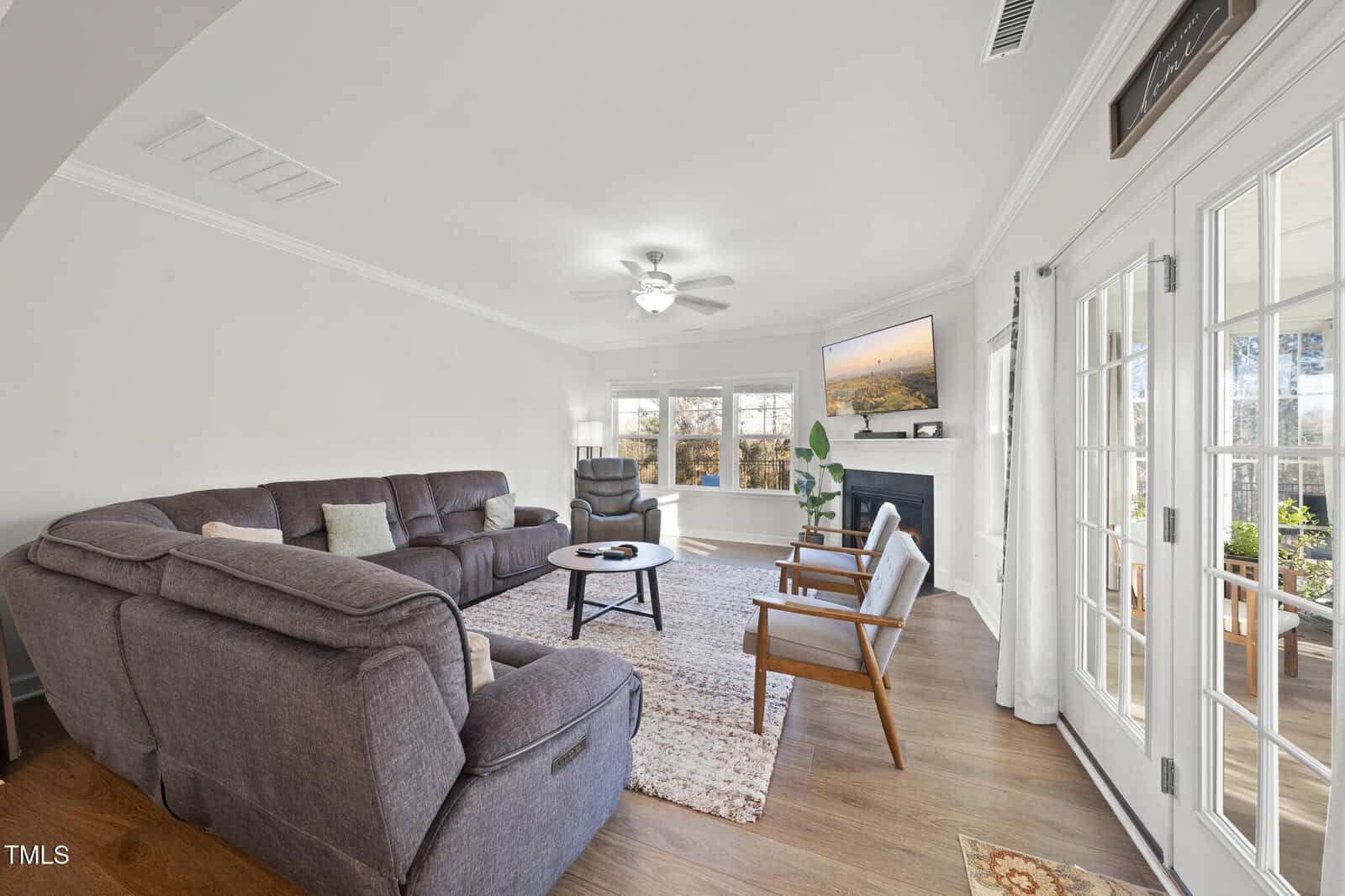 A spacious living room with a large sectional sofa, two armchairs, a coffee table, and a fireplace with a TV above it. Windows and glass doors let in natural light, and there's a ceiling fan on the white ceiling.