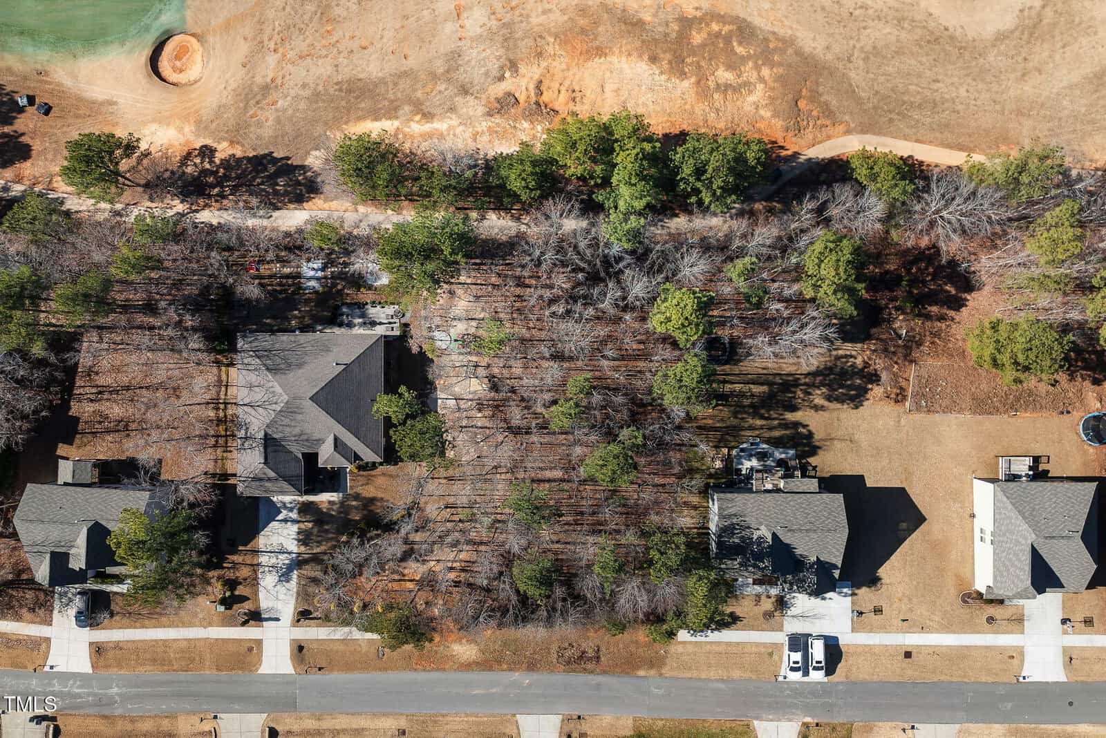 Aerial view of suburban houses with gray roofs, driveways, and lawns; a wooded area separates the homes from a dirt area and golf course in the upper part of the image.