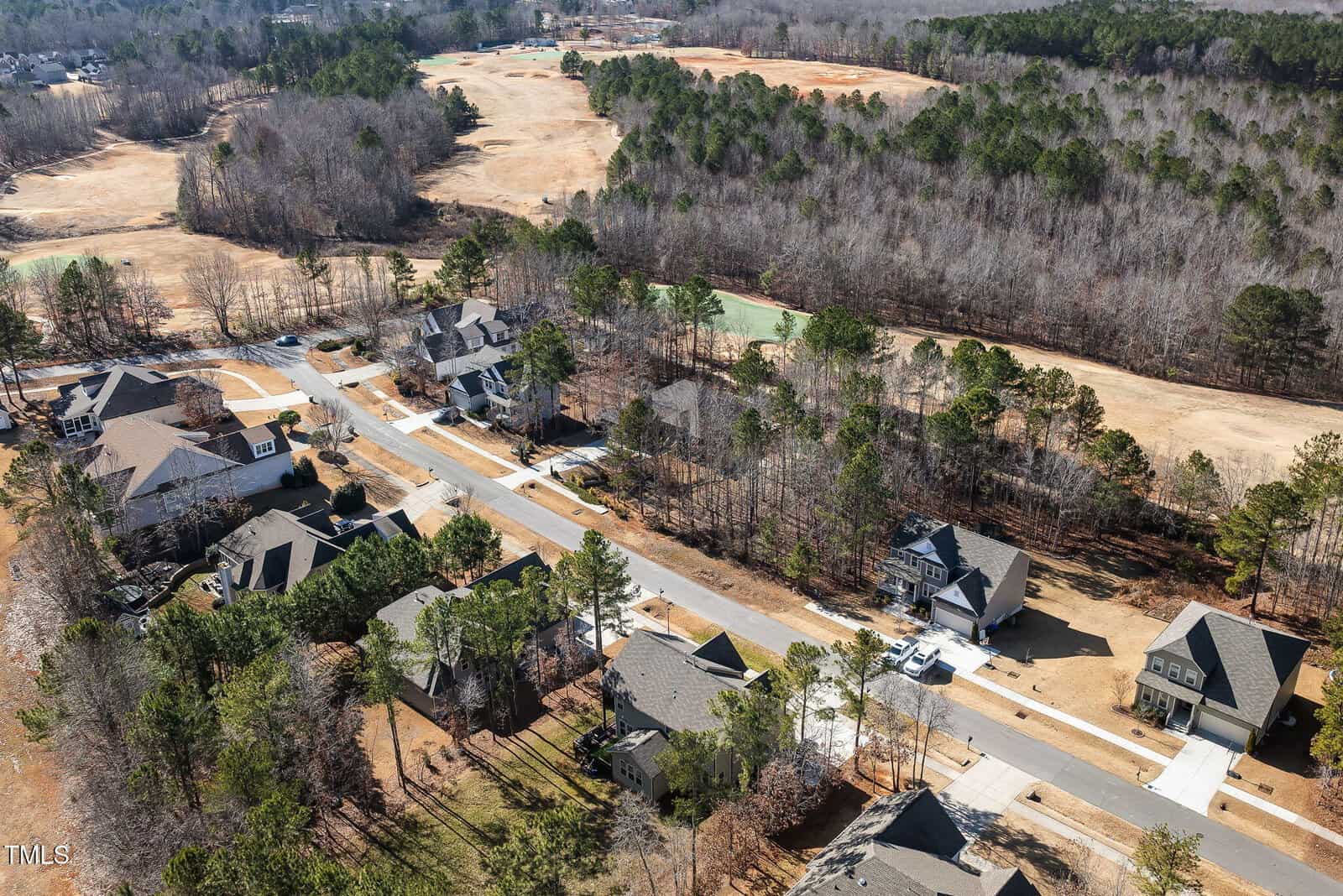 Aerial view of a suburban neighborhood with houses, roads, driveways, and cars. Wooded areas and an open grass field or golf course are visible in the background. Trees surround many properties.