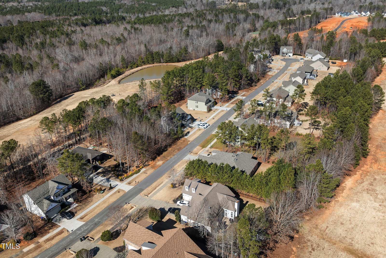 Aerial view of a suburban neighborhood with houses, trees, a road, parked cars, a small pond, and some cleared land. Forested areas surround the development.