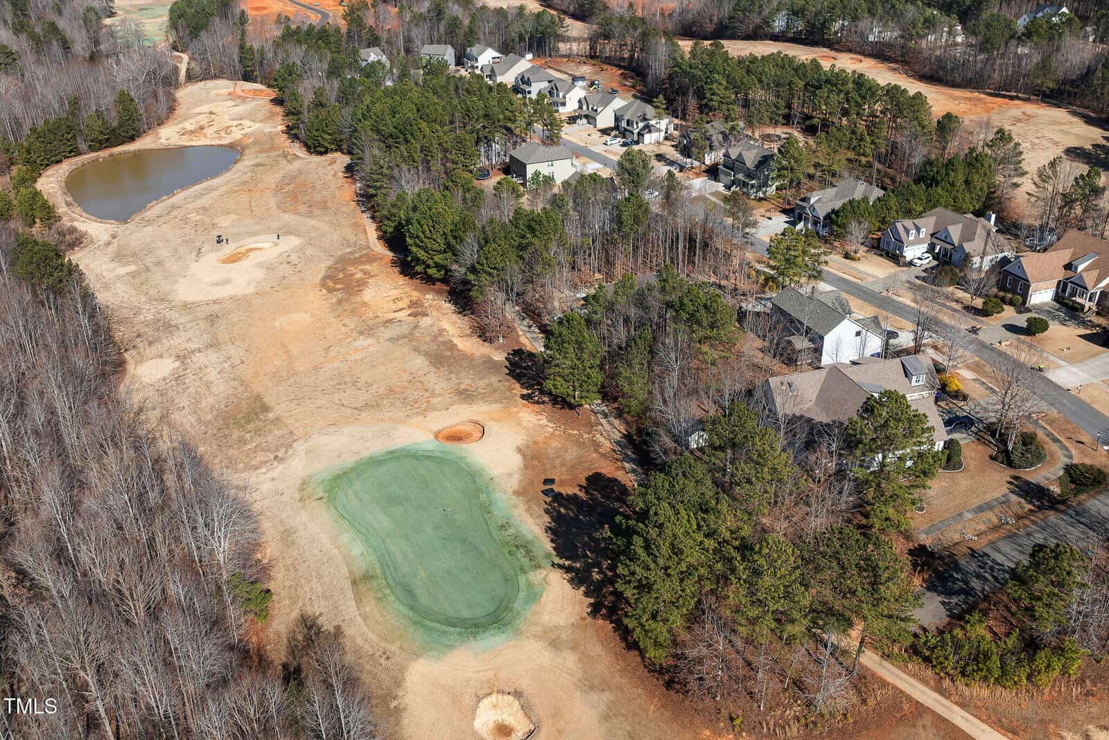 Aerial view of a suburban neighborhood next to a partially dry golf course, with a green, a sand bunker, a pond, and a cluster of houses surrounded by trees.