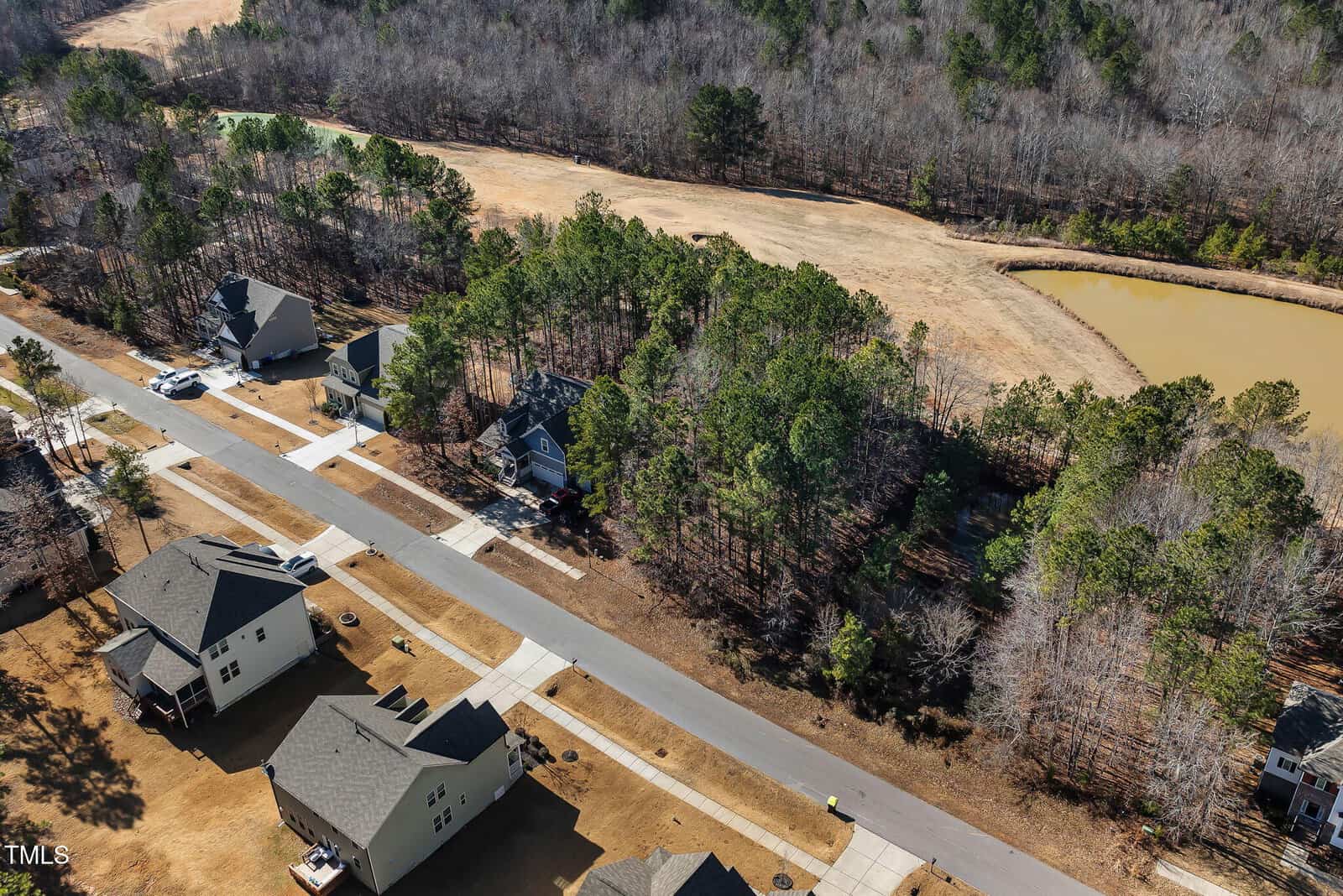 Aerial view of a suburban neighborhood with houses, driveways, and a road. Trees surround the area, and a pond is visible to the right near a cleared field. Some yards appear brown, with scattered cars in driveways.