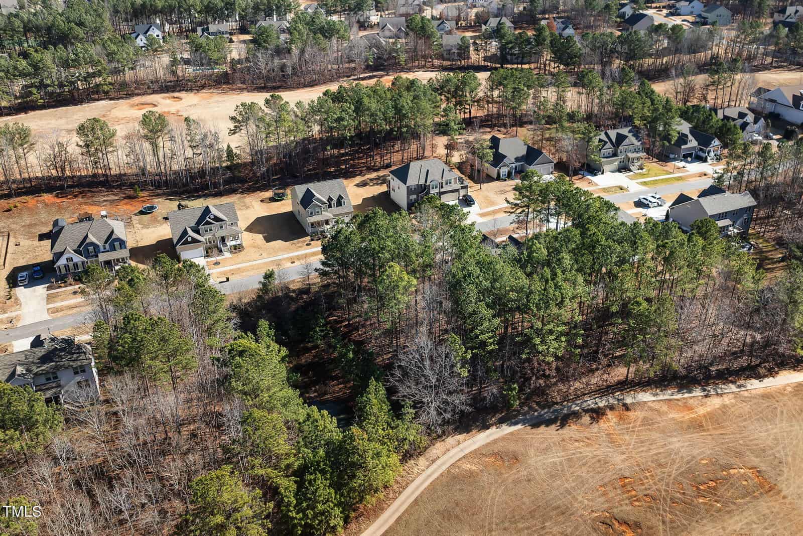 Aerial view of a suburban neighborhood with several houses surrounded by trees. A road runs through the development, and there is a cleared area with red soil and sparse vegetation in the foreground.