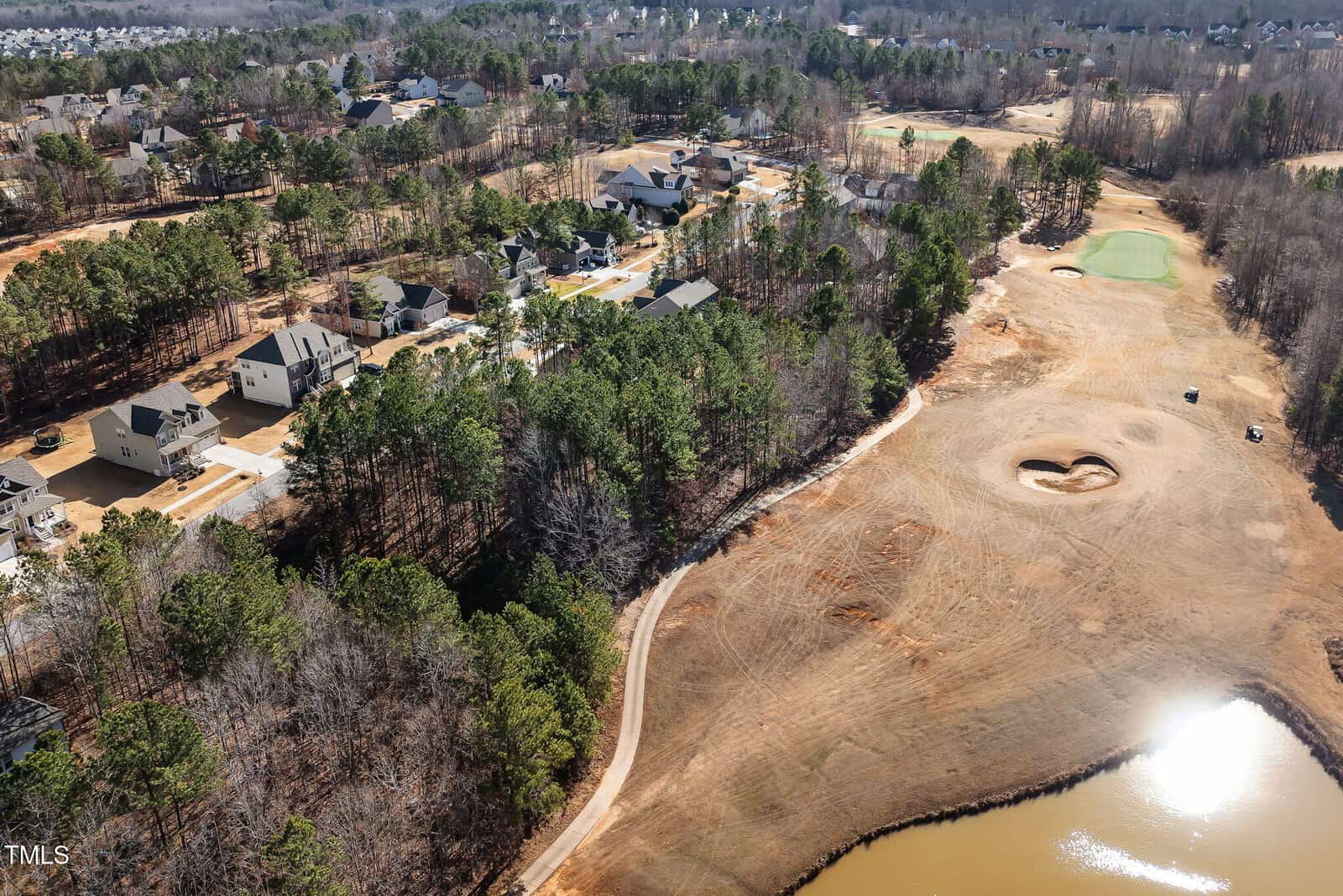 Aerial view of a residential neighborhood adjacent to a golf course with a sand bunker, surrounded by trees and a small pond in the foreground. Houses and streets are visible among wooded areas.