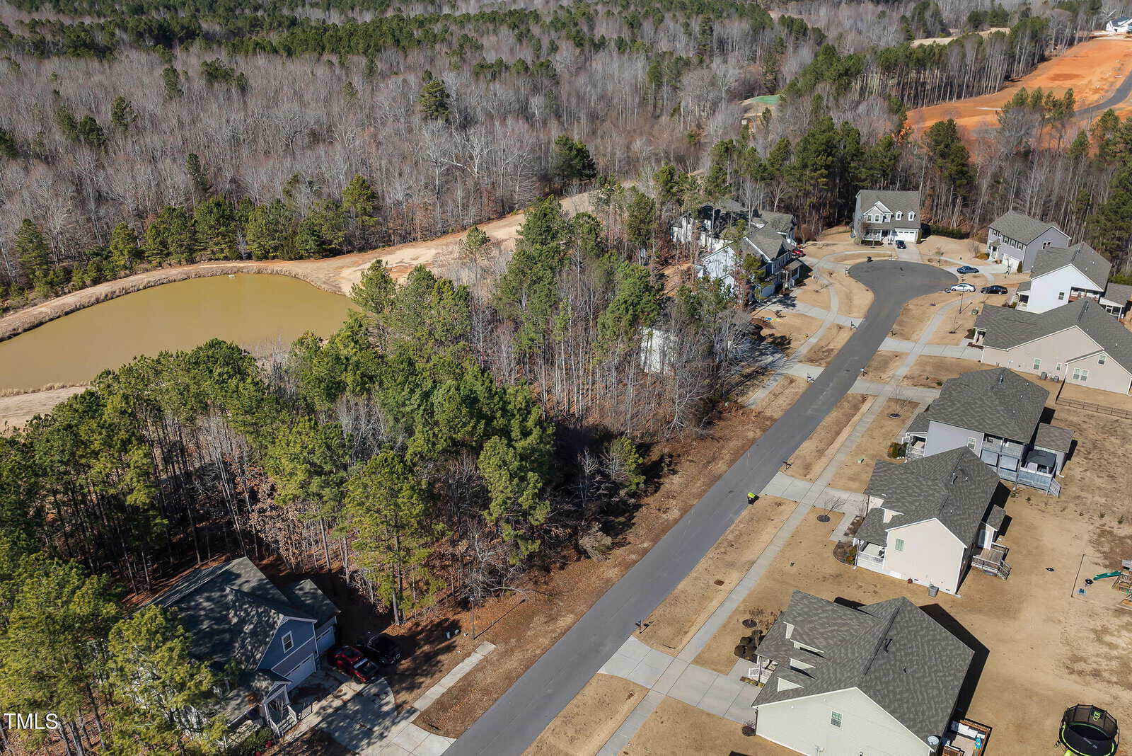 Aerial view of a suburban neighborhood with houses, a curved road, dense trees, and a small pond. The landscape includes a mix of wooded areas and cleared land under development.
