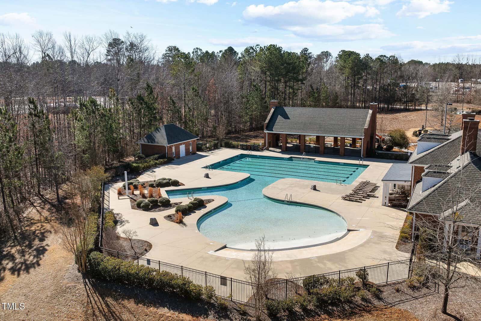 A large outdoor swimming pool with a shallow area, lap lanes, and a covered pavilion is surrounded by lounge chairs and trees. The pool deck is spacious with landscaped planters, and small buildings sit nearby.