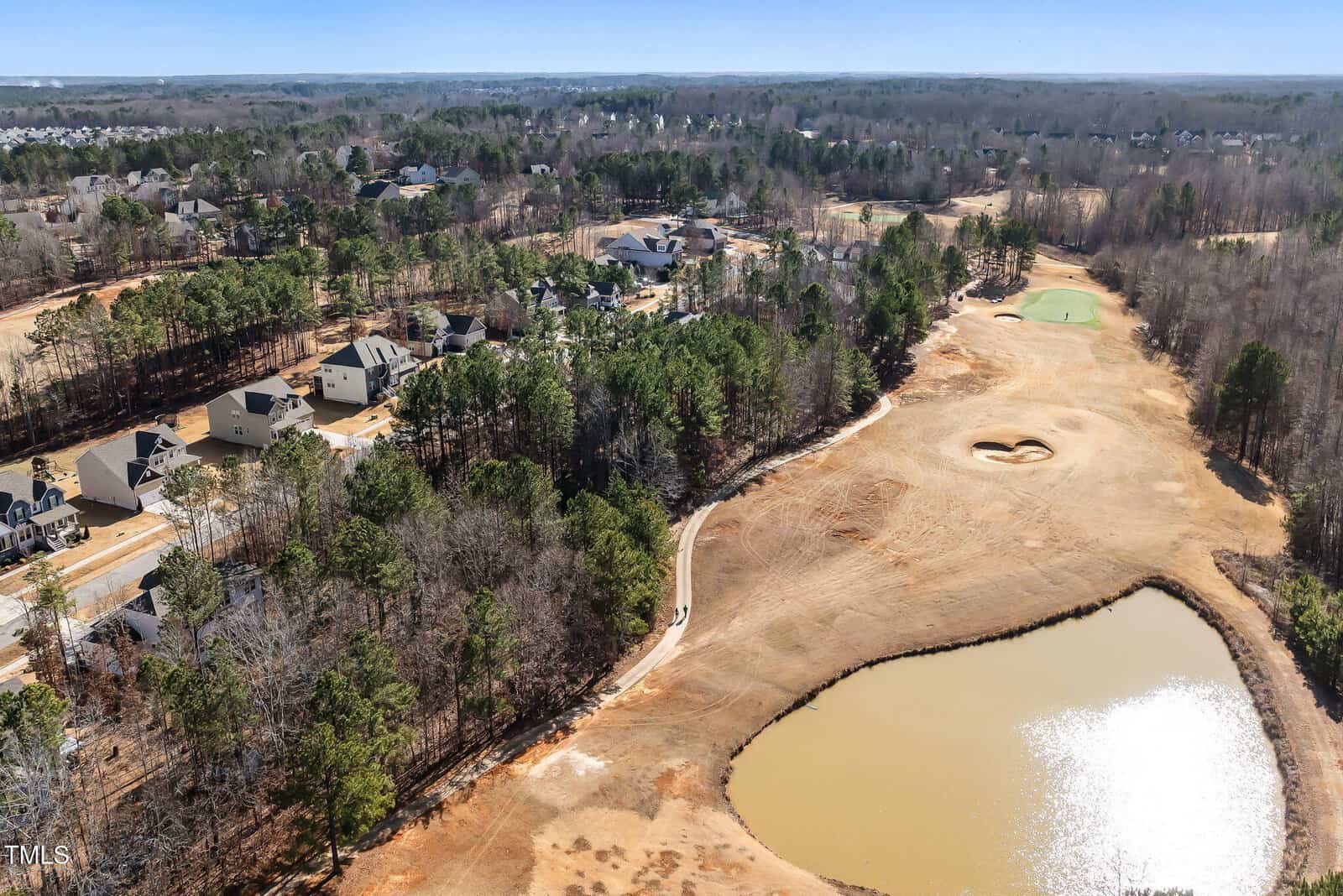 Aerial view of a suburban neighborhood bordering a golf course, with a pond in the foreground, trees separating homes from the course, and the green of a golf hole visible in the distance.