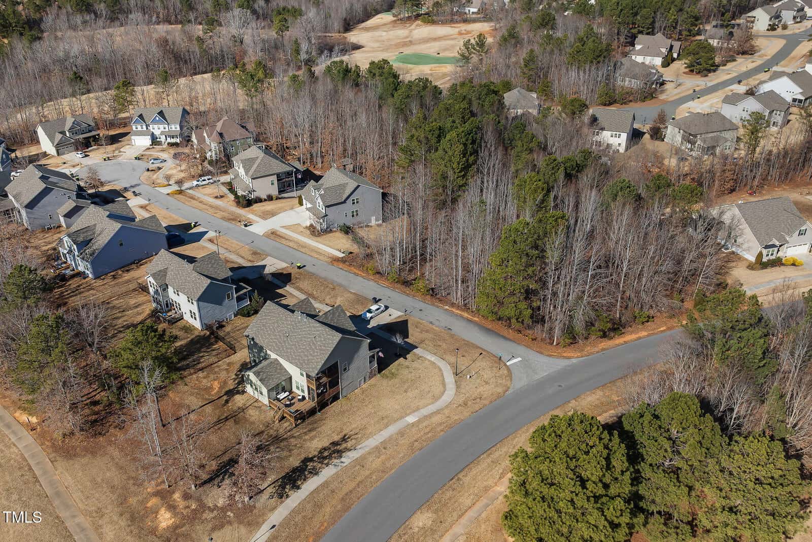 Aerial view of a suburban neighborhood with houses, driveways, and roads surrounded by leafless trees and patches of brown grass, likely during late fall or winter.