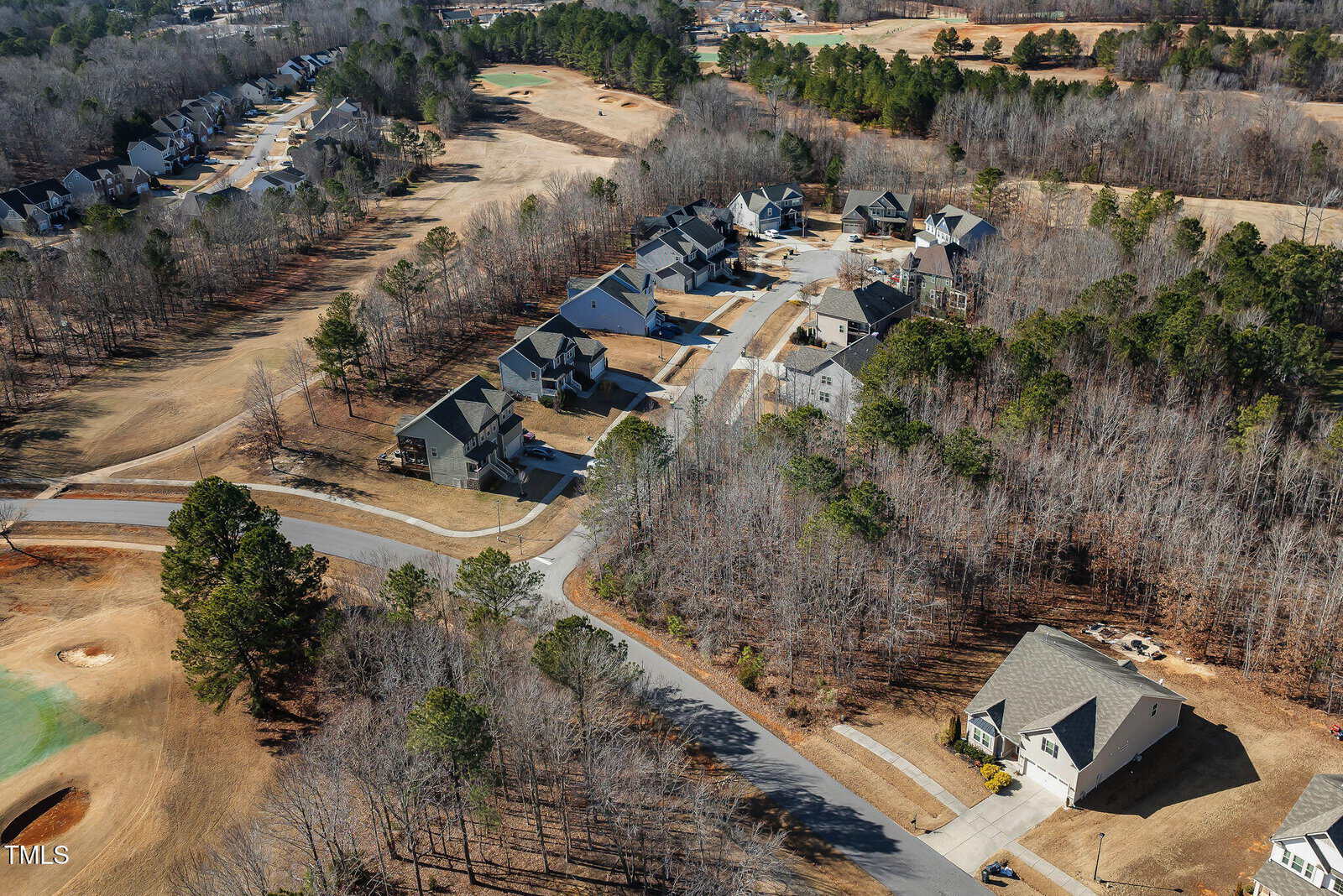 Aerial view of a suburban neighborhood with houses, roads, leafless trees, and a golf course. The area has both wooded sections and open grassy spaces, with homes grouped along winding streets.