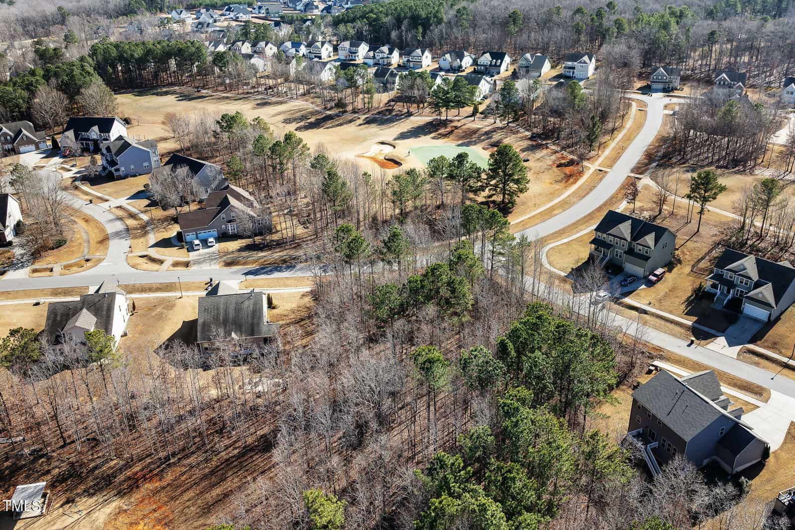 Aerial view of a suburban neighborhood with houses, winding roads, leafless trees, grassy areas, and a golf course. The homes are spaced apart, and the scene shows a mix of woodland and cleared land.