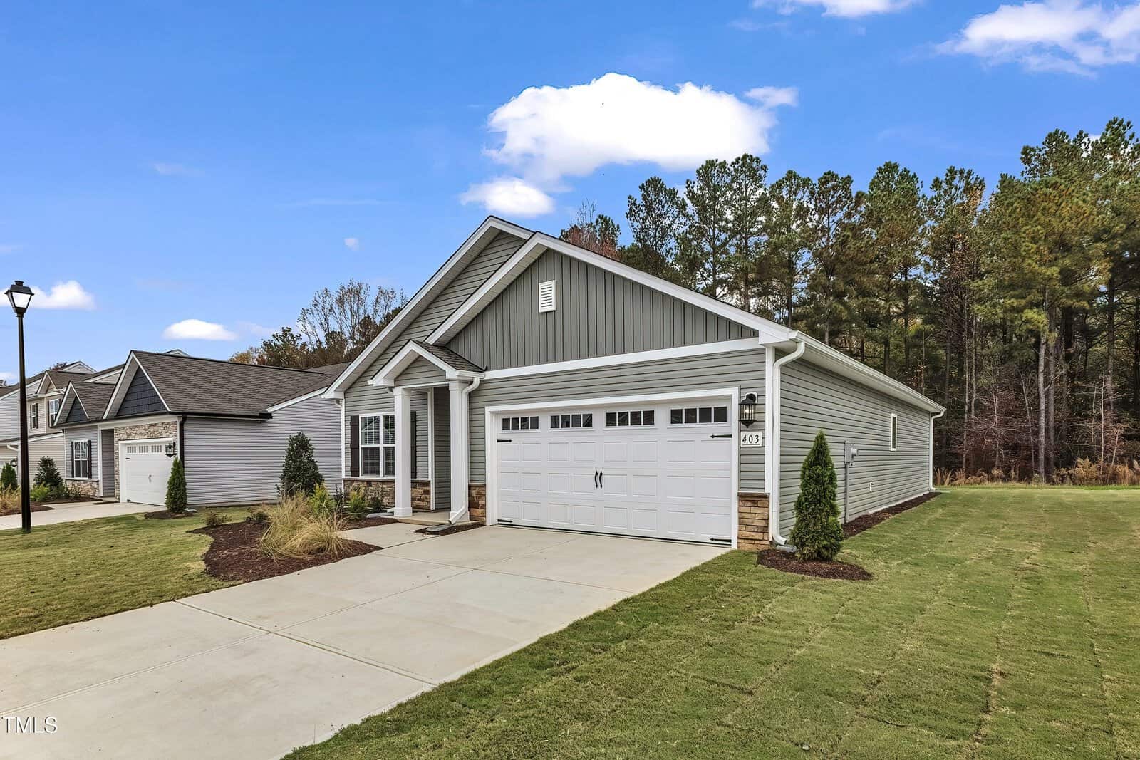 Single-story gray house with white trim and a two-car garage, set on a neatly landscaped lawn with a concrete driveway. Trees are visible in the background under a partly cloudy sky.