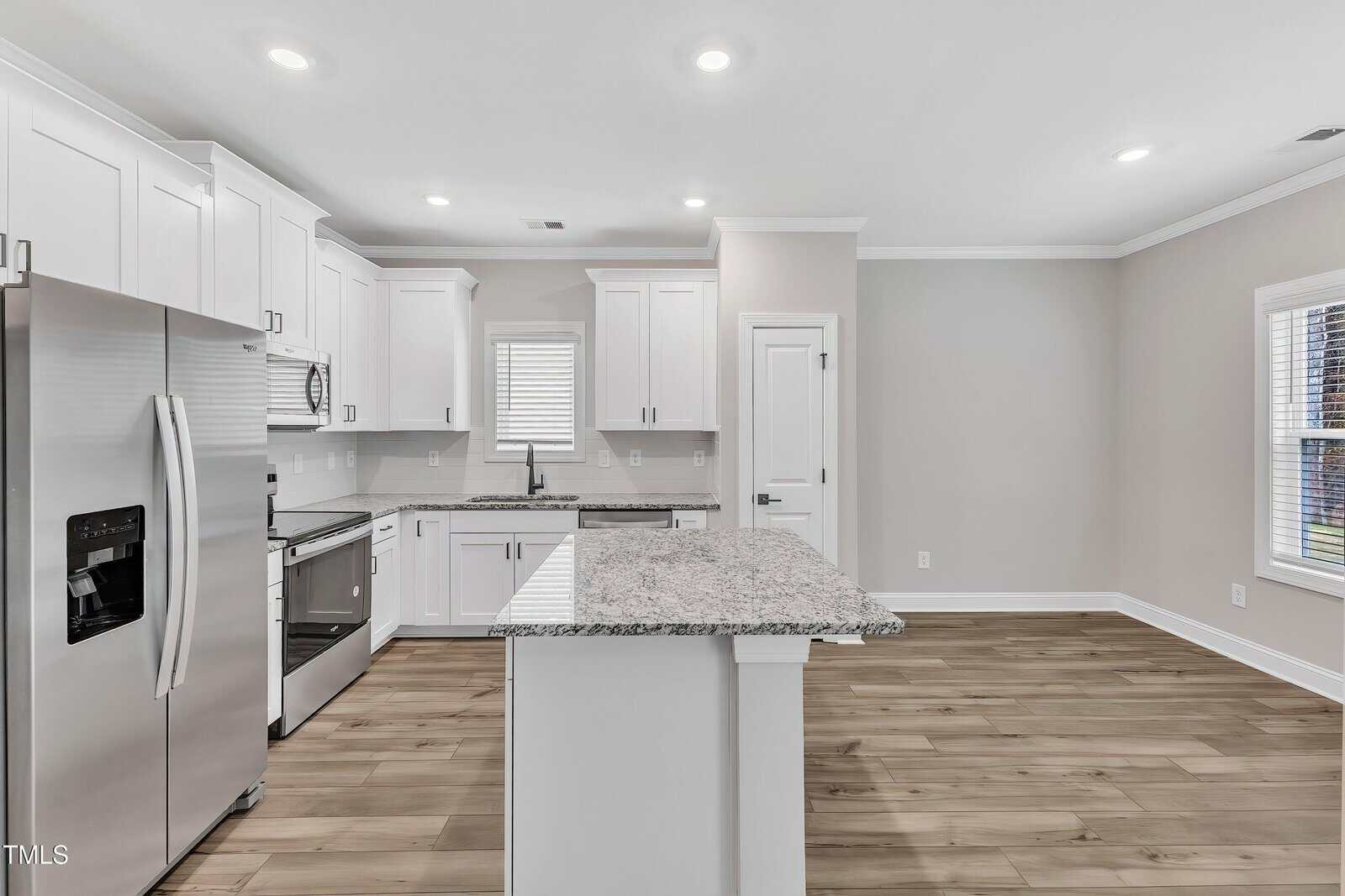 Modern kitchen with white cabinets, stainless steel appliances, a center island with a granite countertop, light wood flooring, and recessed ceiling lights. A window and a door are visible on the back wall.