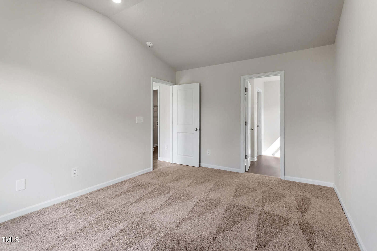 Empty room with light beige carpet and neutral-colored walls. There are two white doors; one is open, leading to a hallway with visible light, and the other is closed. The ceiling is plain with a small light fixture.