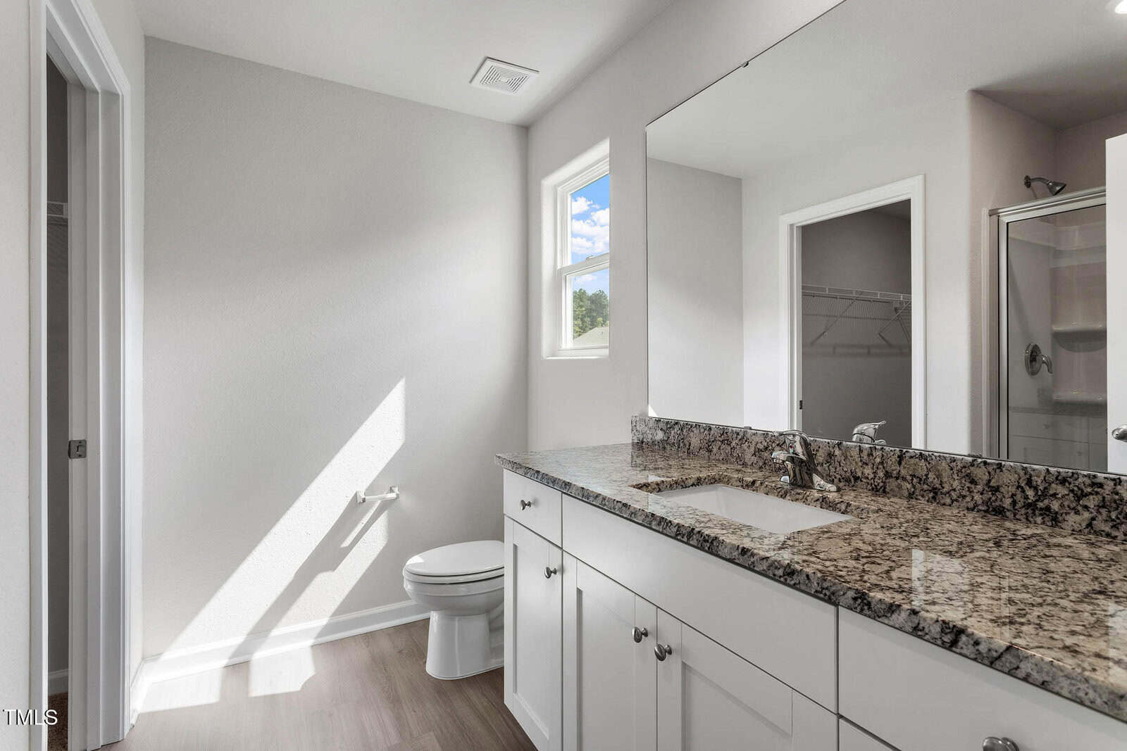 A modern bathroom features a large mirror above a granite countertop with a sink. There is a toilet next to a window letting in natural light. The floor is wooden, and a shower stall is partially visible in the background.