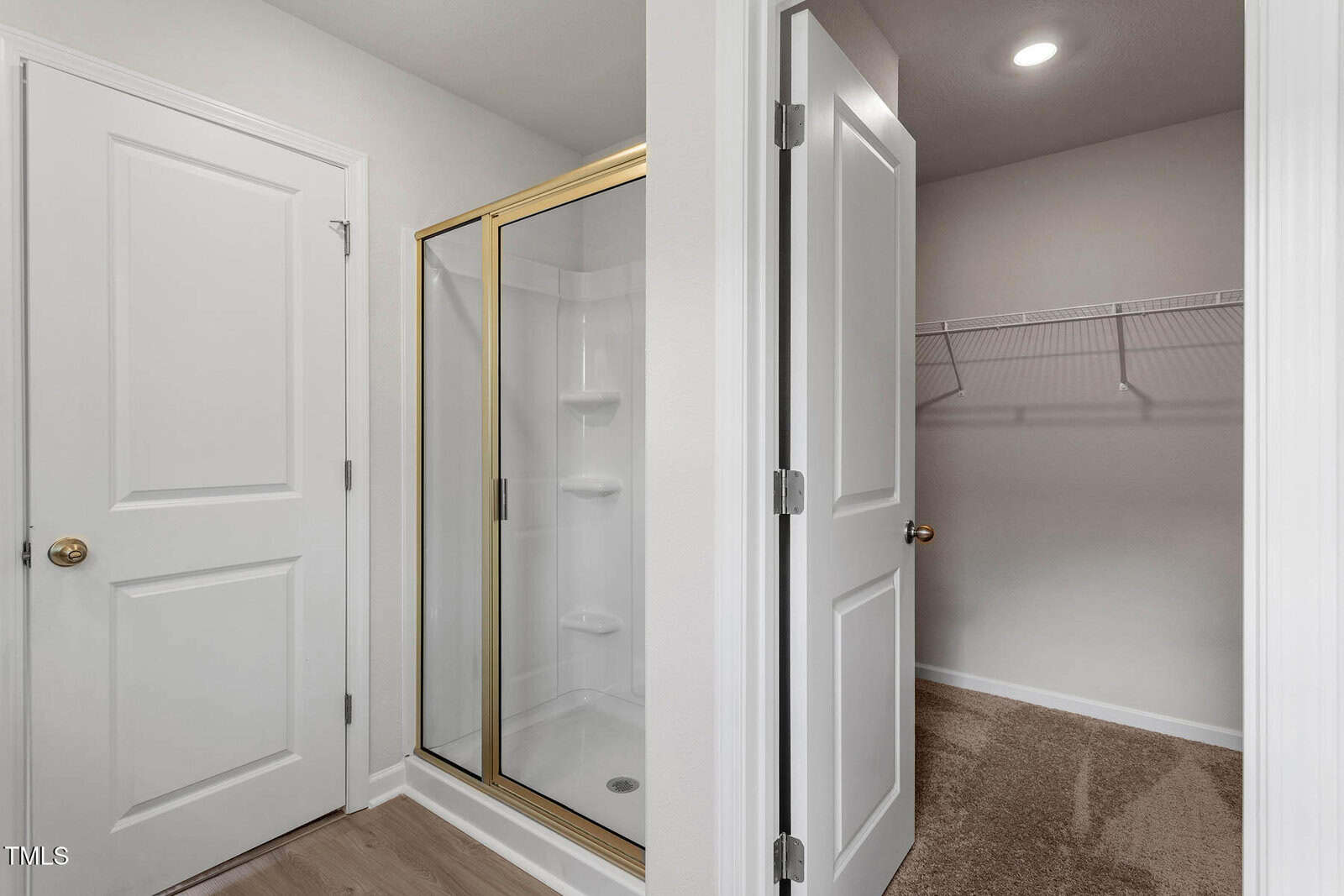 A bathroom with a glass-enclosed shower on the left, featuring white walls and gold trim. To the right is an open door revealing a small carpeted walk-in closet with a wire shelf. Both spaces have white doors and beige-colored walls.