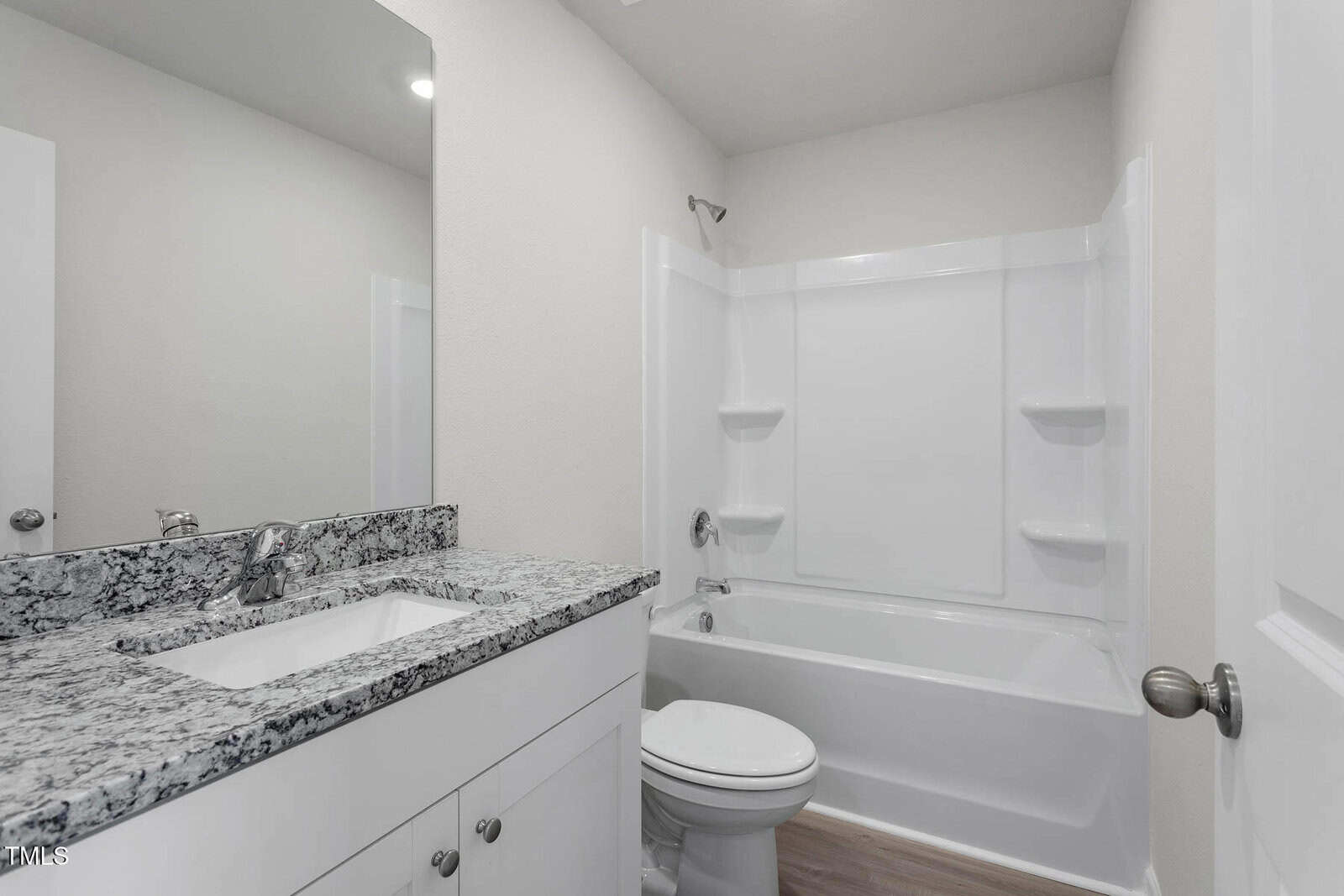 A bathroom with a white bathtub and shower, granite countertop sink, and a toilet. The walls are light-colored, and the floor is wood-patterned. A mirror is above the sink, reflecting the light and ceiling.