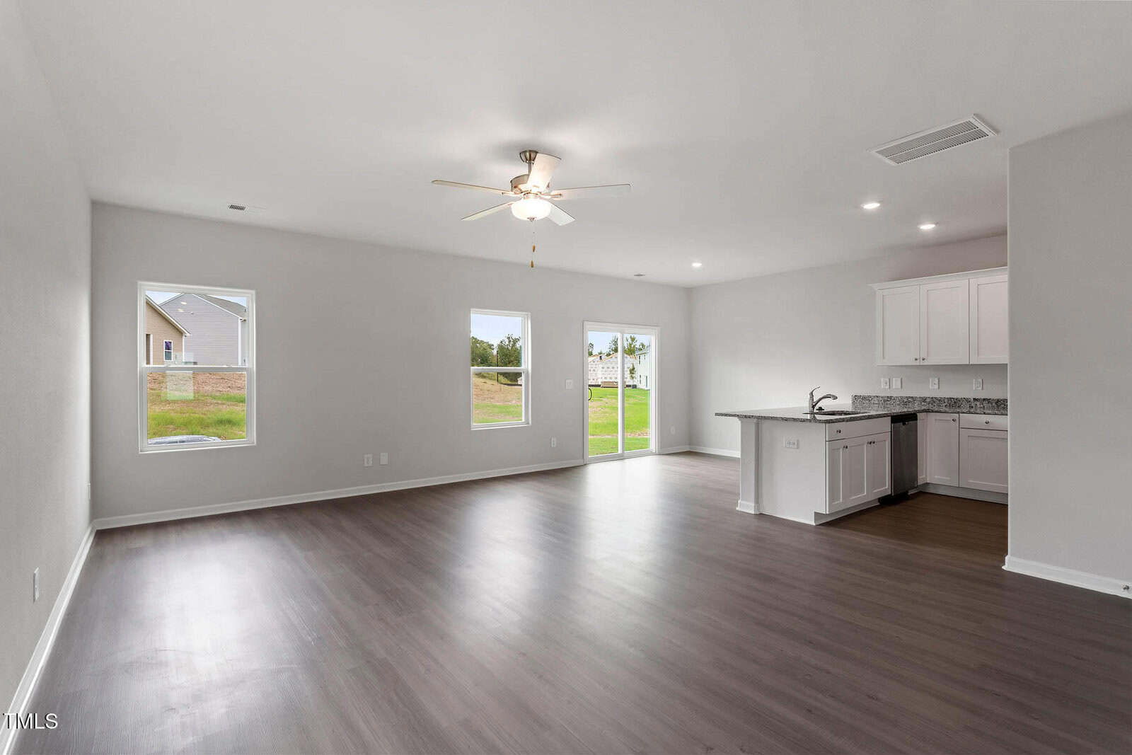 Spacious living area with light-colored walls and wooden floors. A ceiling fan hangs above. Two windows and a glass door provide natural light. The open kitchen features white cabinets and dark countertops, with a view of a green lawn outside.