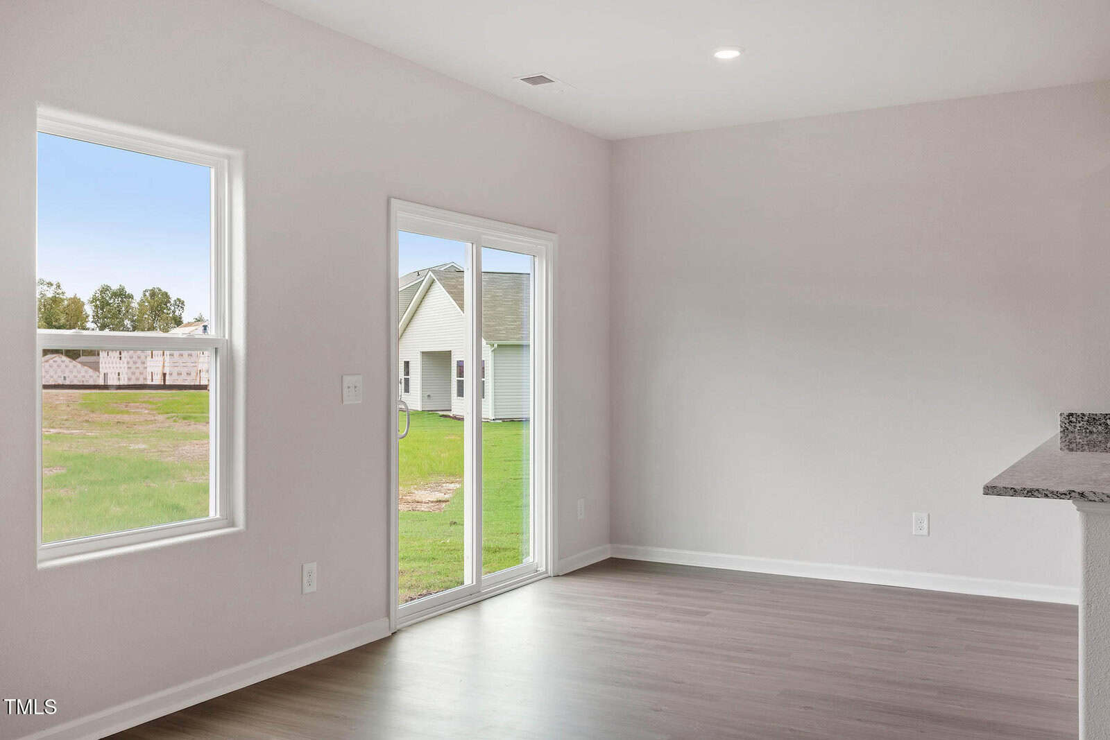 A bright, empty room with light-colored walls and wood flooring. There is a large window and sliding glass doors leading outside. A small portion of a kitchen counter is visible on the right.