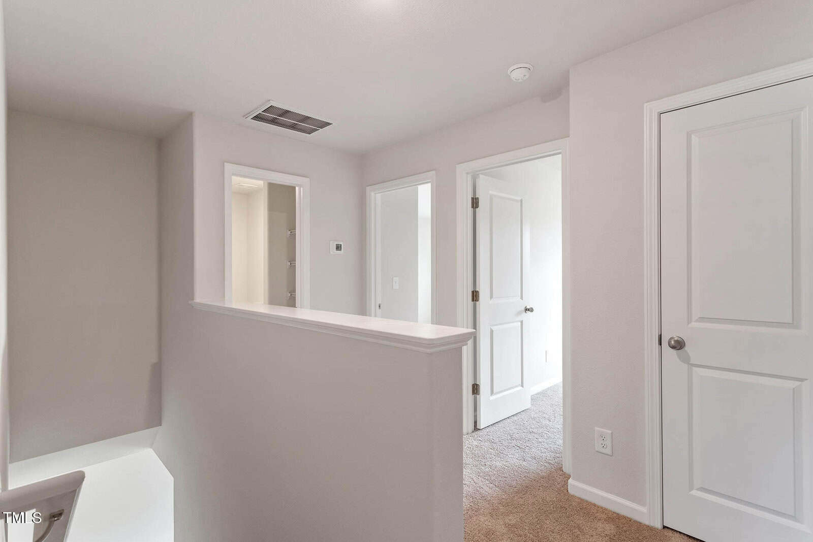 Hallway with beige walls and carpeted floor. There is a white door on the right, an open door leading to another room, and an additional doorway on the left. A half-wall is shown in the foreground. Ceiling lights illuminate the space.