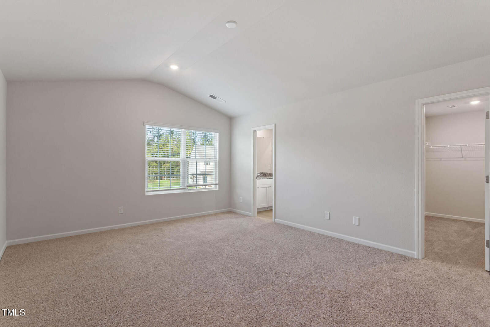 Empty carpeted room with a large window on the left wall. The walls are painted light gray. There is a doorway on the right leading to a walk-in closet, and another doorway to a small bathroom. Ceiling lights are on.