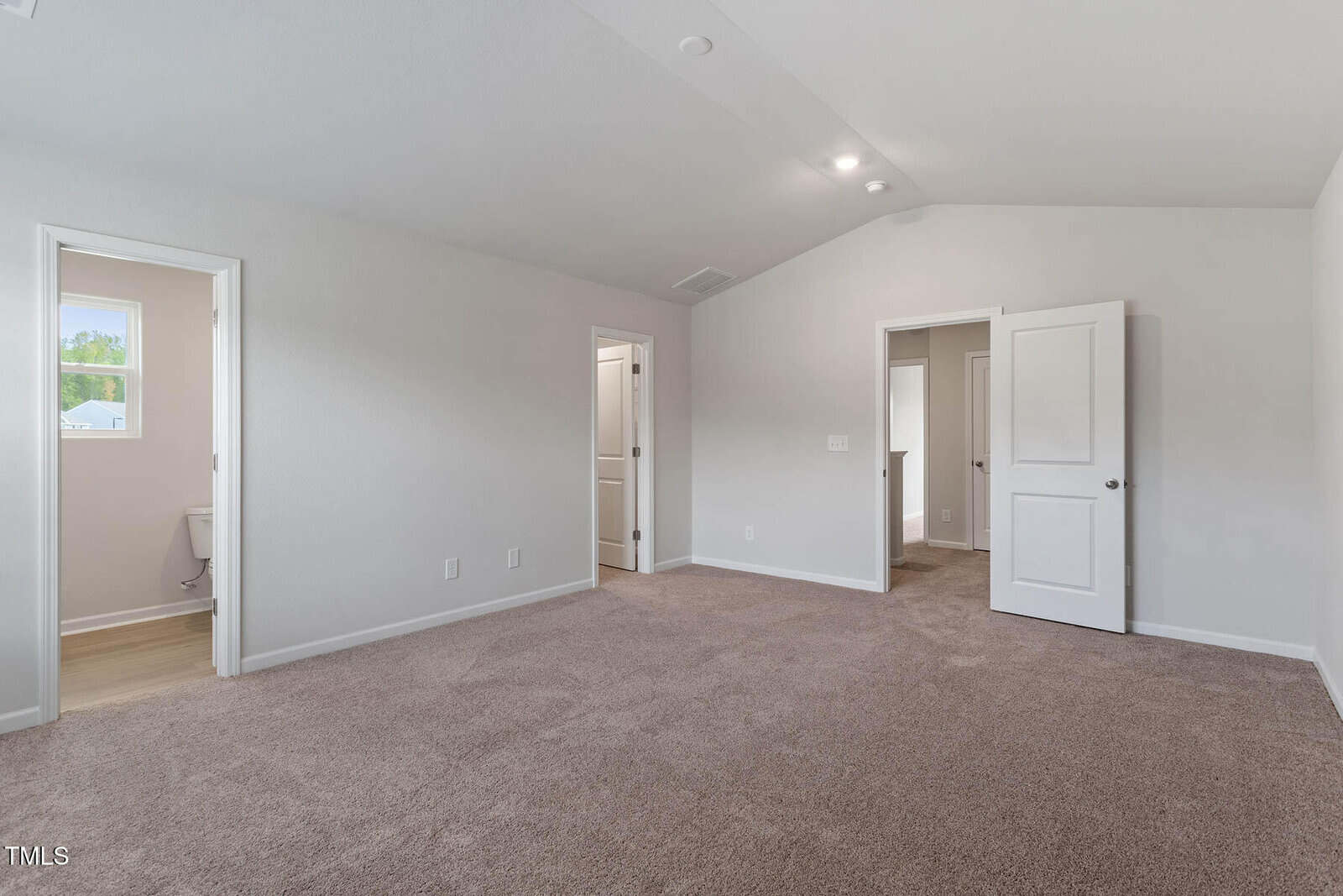 Empty beige-carpeted bedroom with white walls and a vaulted ceiling. Two open doorways leading to adjacent rooms and a bathroom. Natural light from a window in the bathroom illuminates the space.